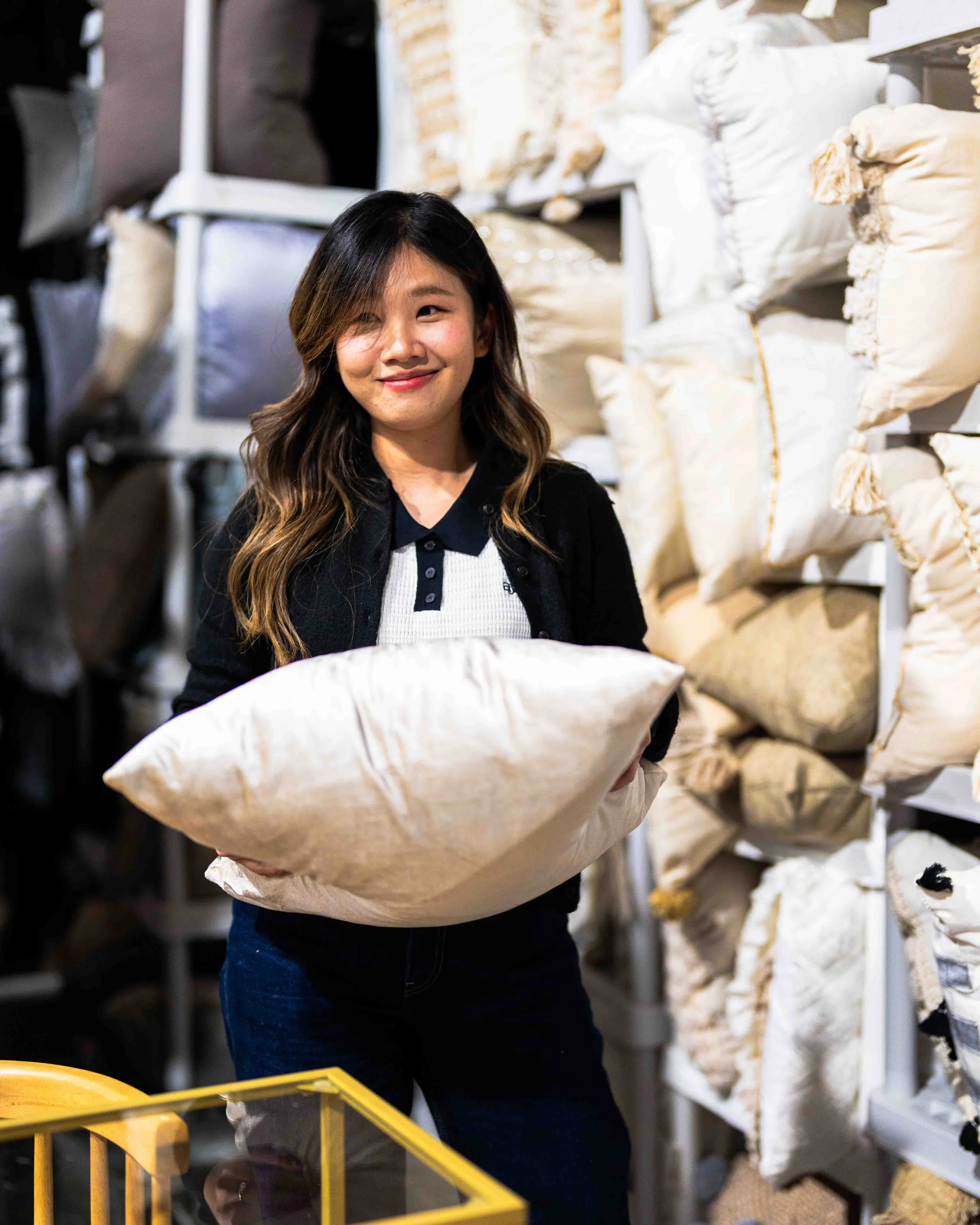 A young woman in a black jacket and striped shirt holding a large white pillow in a store aisle with shelves filled with cushions and fabric in the background.