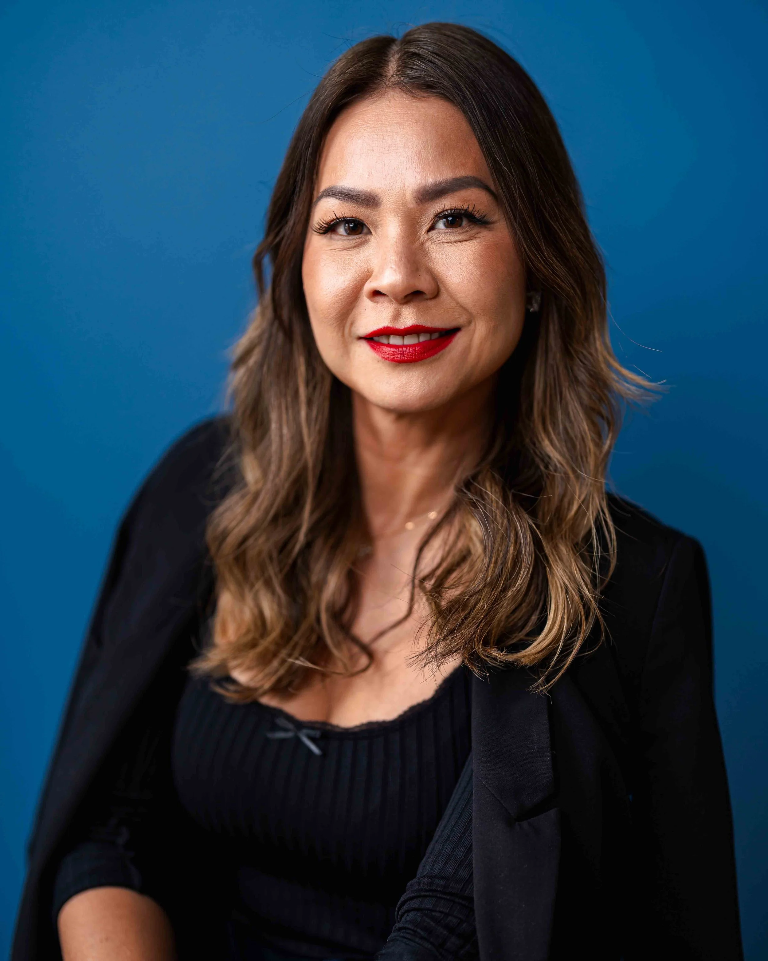 Close-up portrait of a smiling woman with long wavy brown hair, wearing a black top and red lipstick, against a blue background.