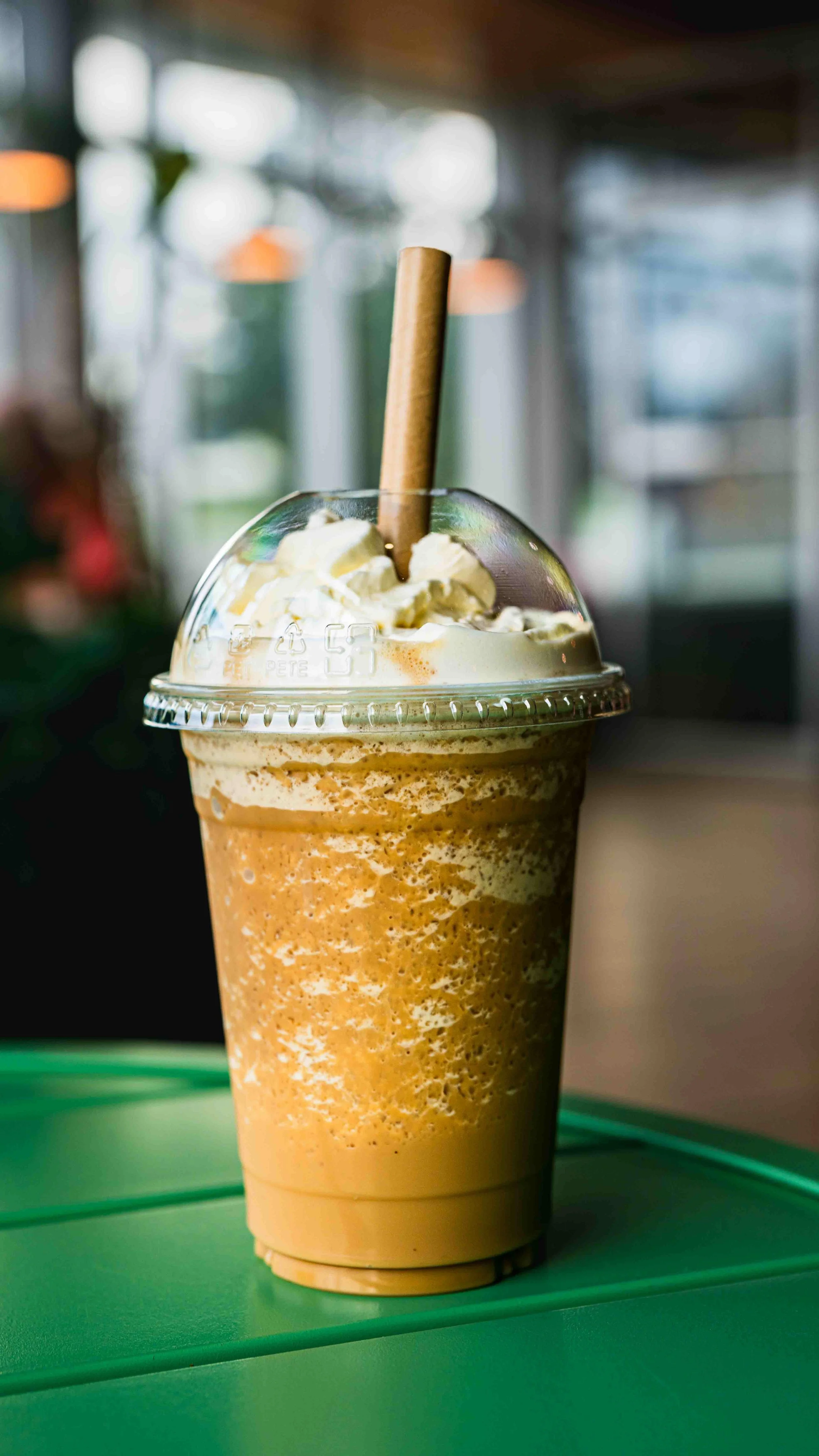 A blended coffee drink topped with whipped cream and a wafer cookie in a clear plastic cup with a dome lid and a brown paper straw, sitting on a green tray.