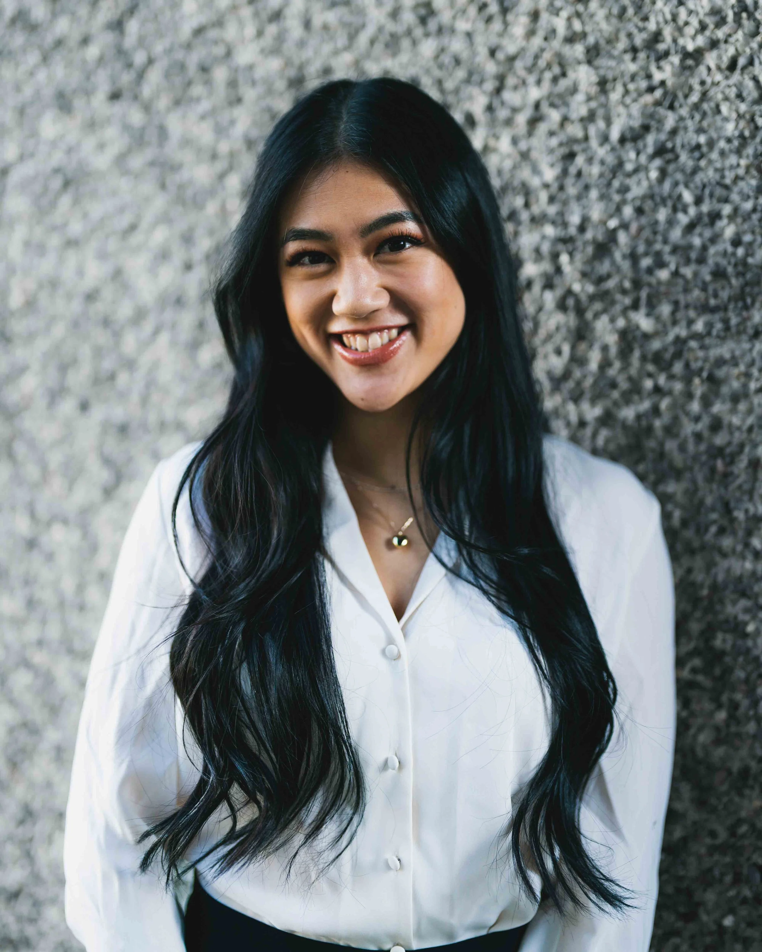 A young woman with long black hair and a radiant smile standing against a textured gray background, wearing a white button-up shirt.