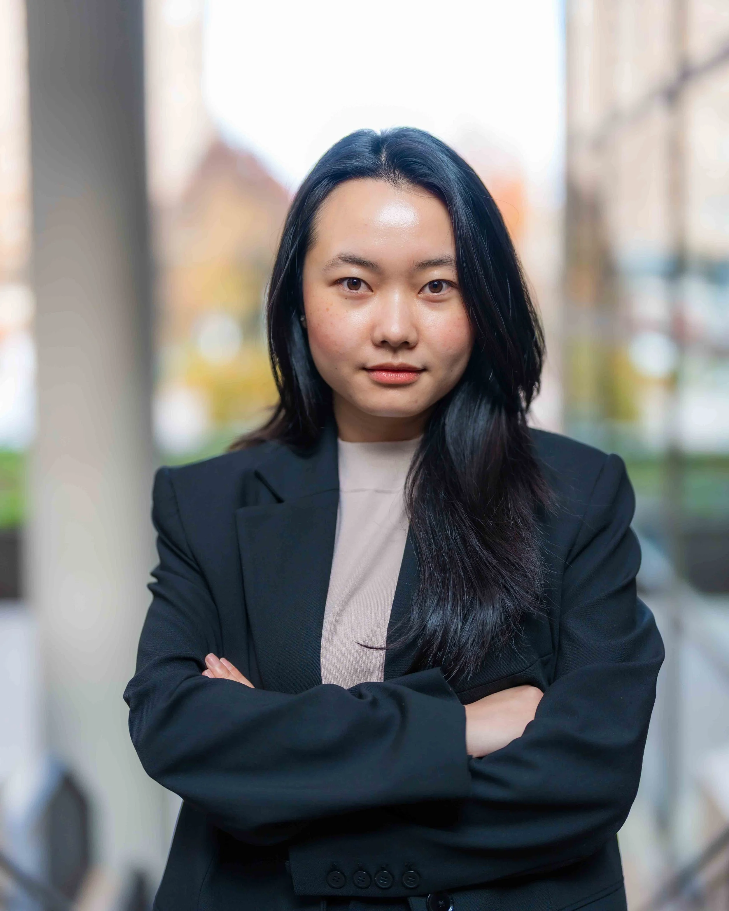 Portrait of a woman with black hair, wearing a black blazer and a beige top, standing with arms crossed outdoors, blurred city background.