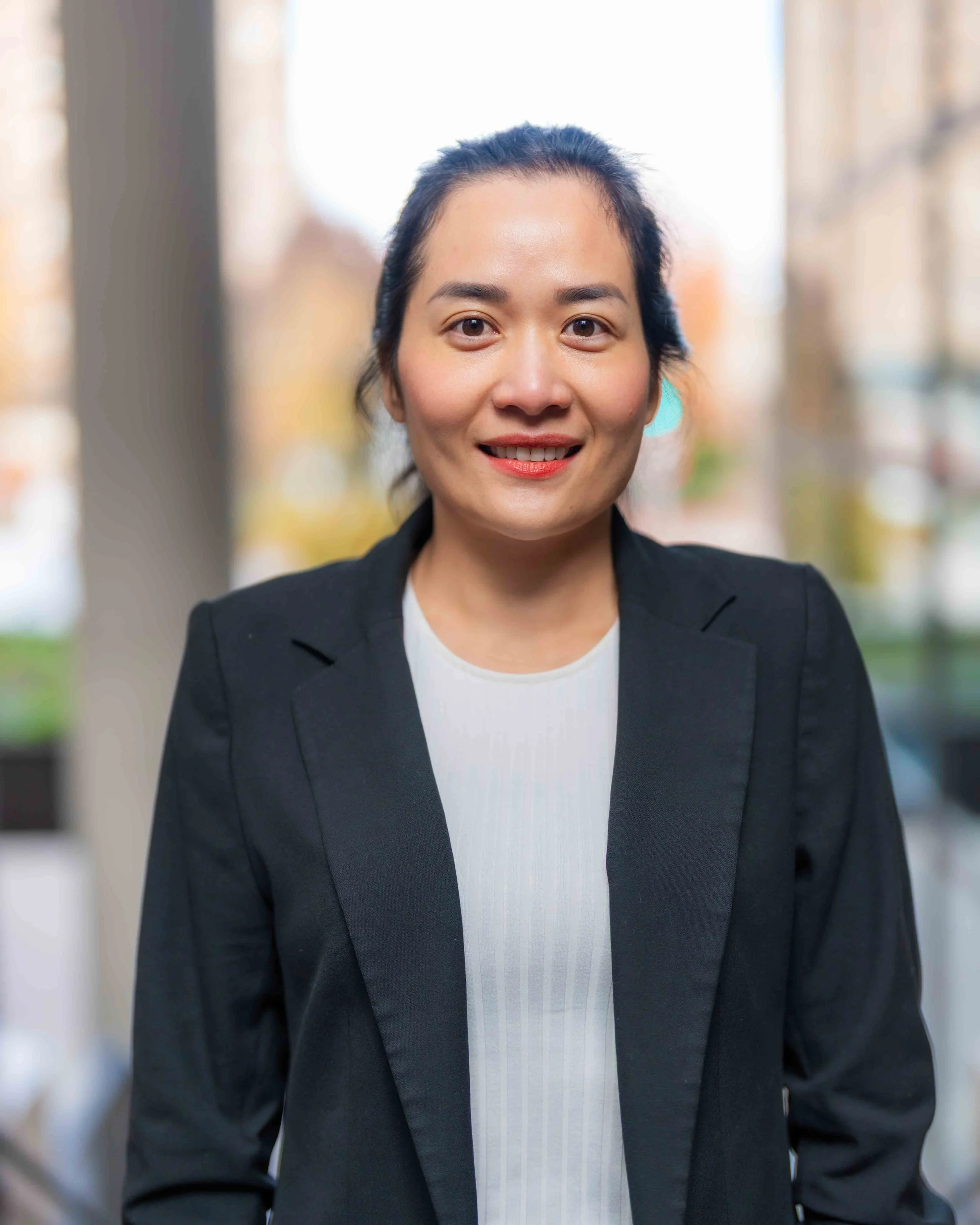 Close-up of an Asian woman with dark hair pulled back, smiling, wearing a black blazer and white shirt, with a blurred outdoors background.