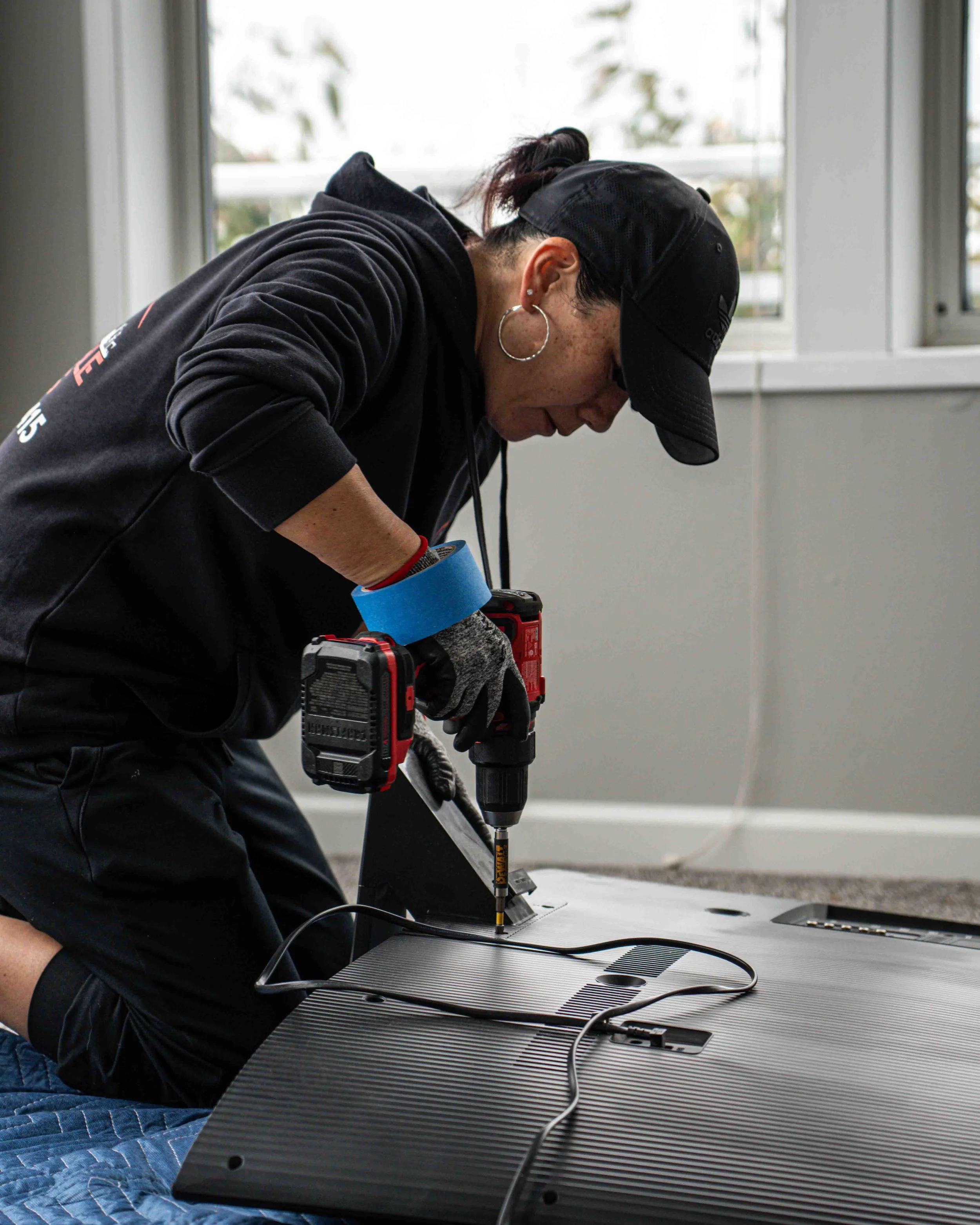 Person using a power drill to assemble or repair a flat-screen monitor on a worktable in a well-lit room.