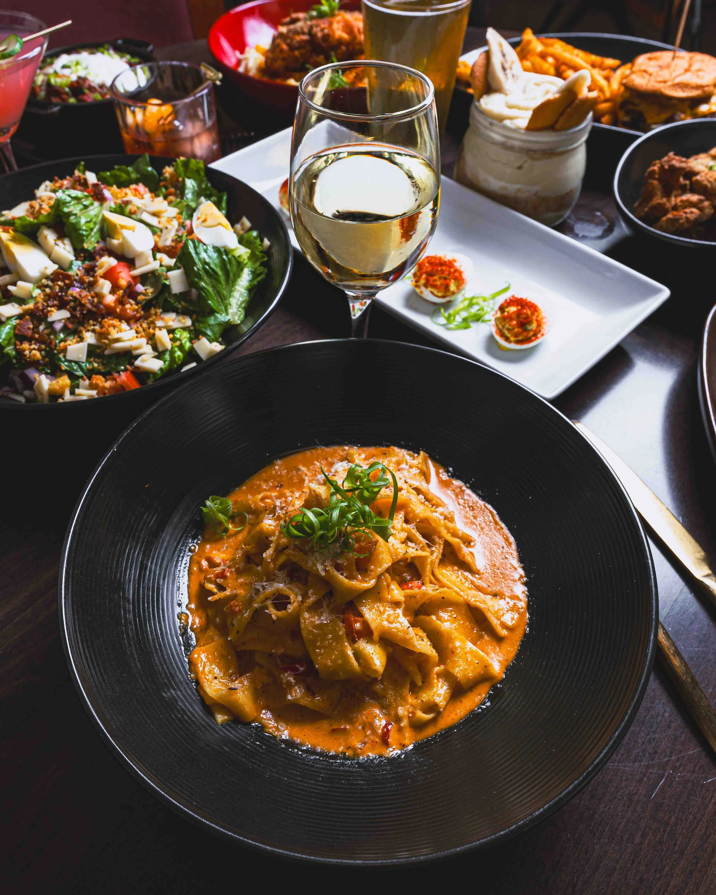 A plate of pasta covered in a creamy tomato sauce and garnished with green onions, surrounded by various dishes including salad, tacos, fried chicken, and two glasses of white wine on a dark table.