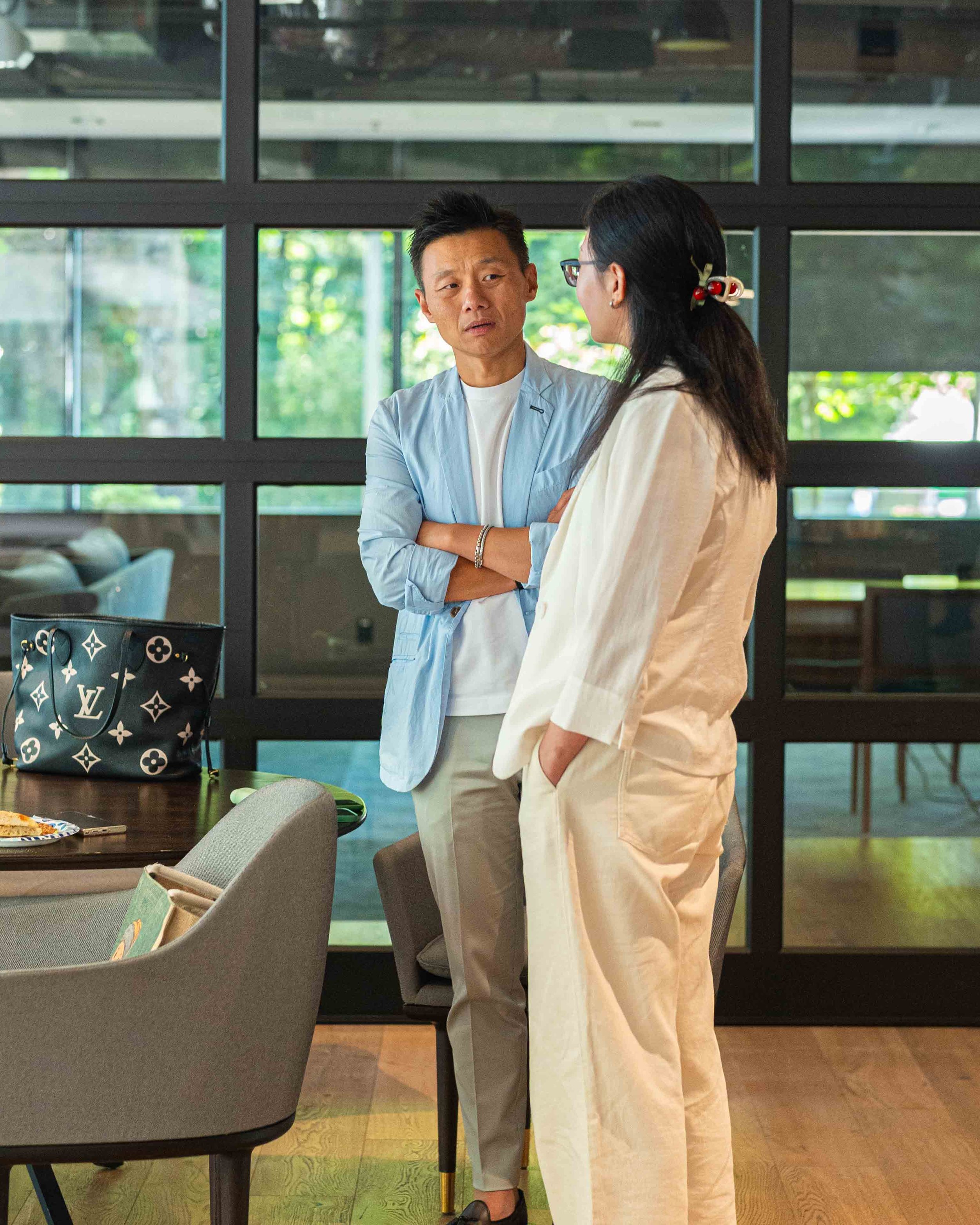 Two people, a man and a woman, are engaged in a conversation indoors, with large windows showing greenery outside.