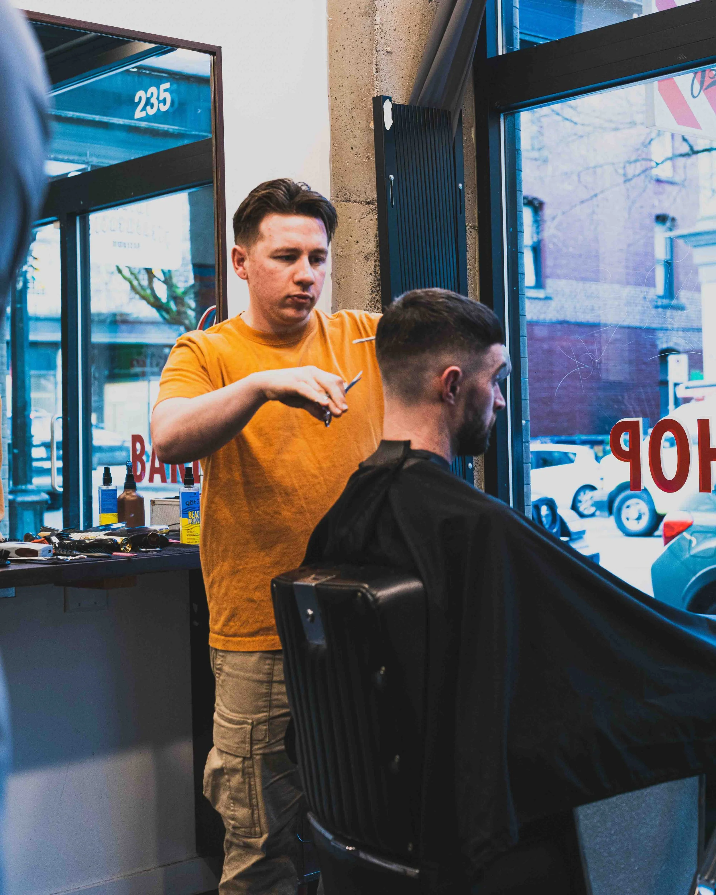 A barber shop scene showing a barber in an orange shirt cutting a customer's hair. The customer is seated in a barber chair with a black cape. The background has a window with a view of parked cars outside, and hair styling tools and products are on 