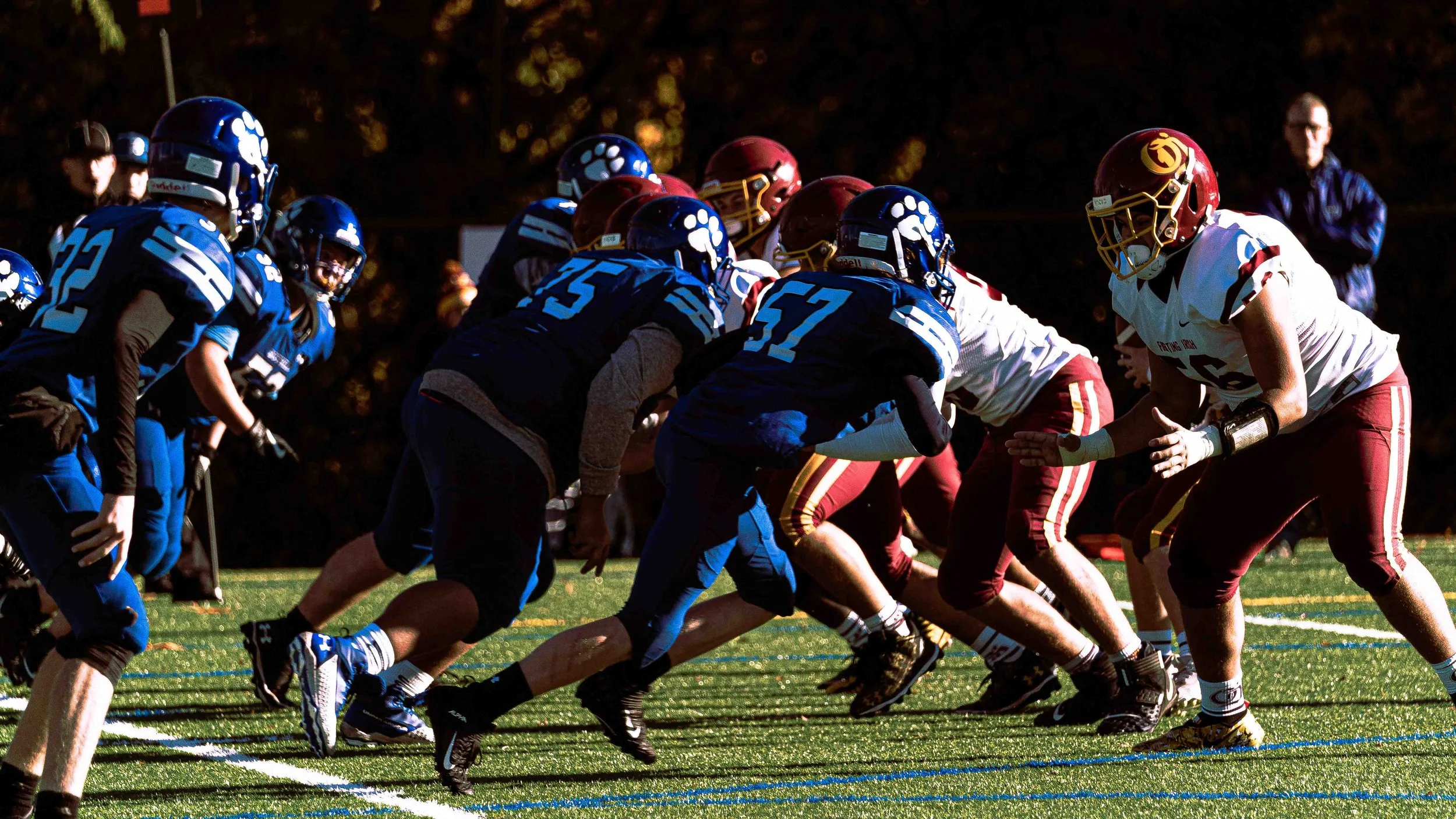 A high school football game with players lined up at the line of scrimmage, wearing blue and white or maroon and yellow uniforms, on a football field during the day.