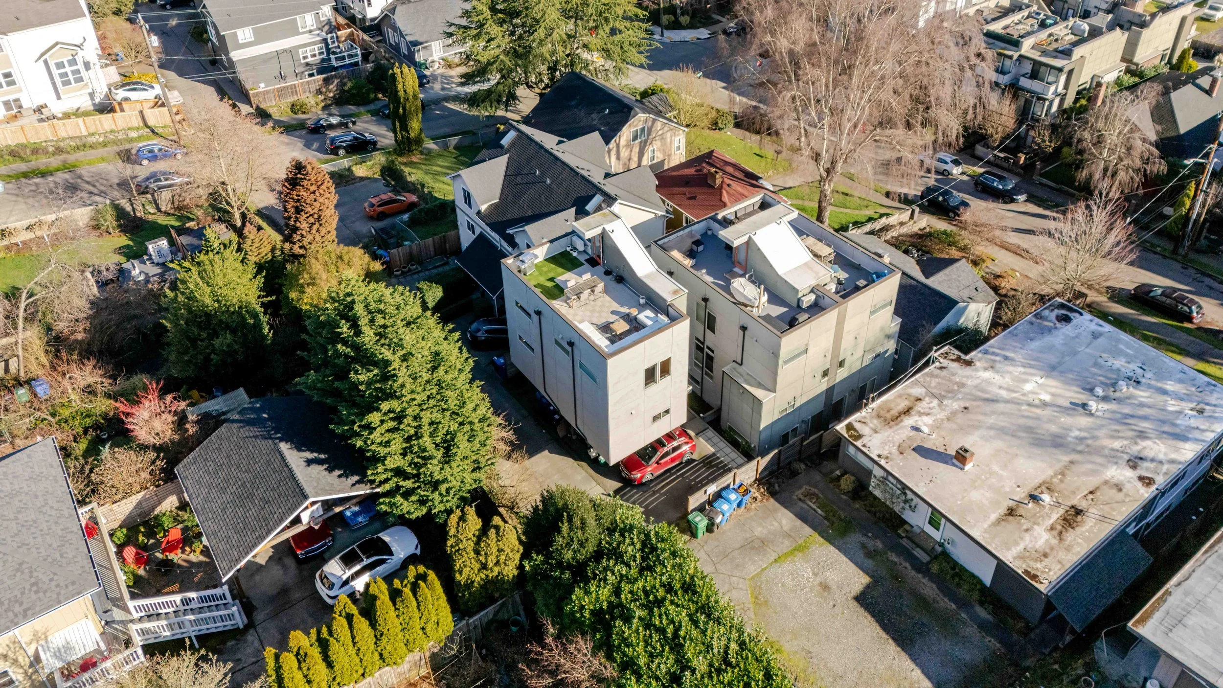 An aerial view of a residential neighborhood featuring a modern multi-story building with rooftop seating, parked cars, leafless and green trees, and surrounding houses, some with fences and driveways.