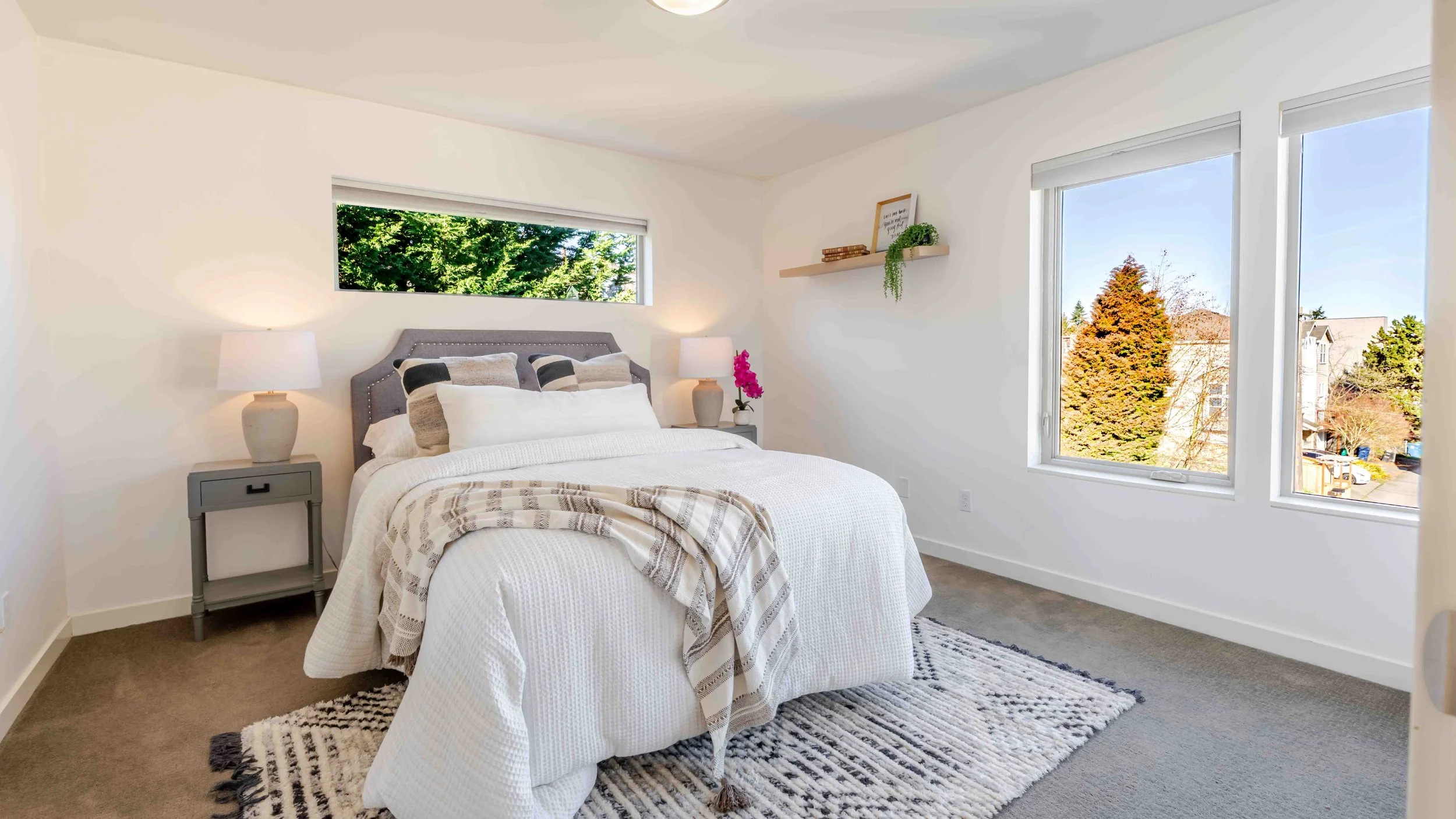 A bedroom with a bed, two nightstands with lamps, a window with a view of trees and houses, and a ceiling light.