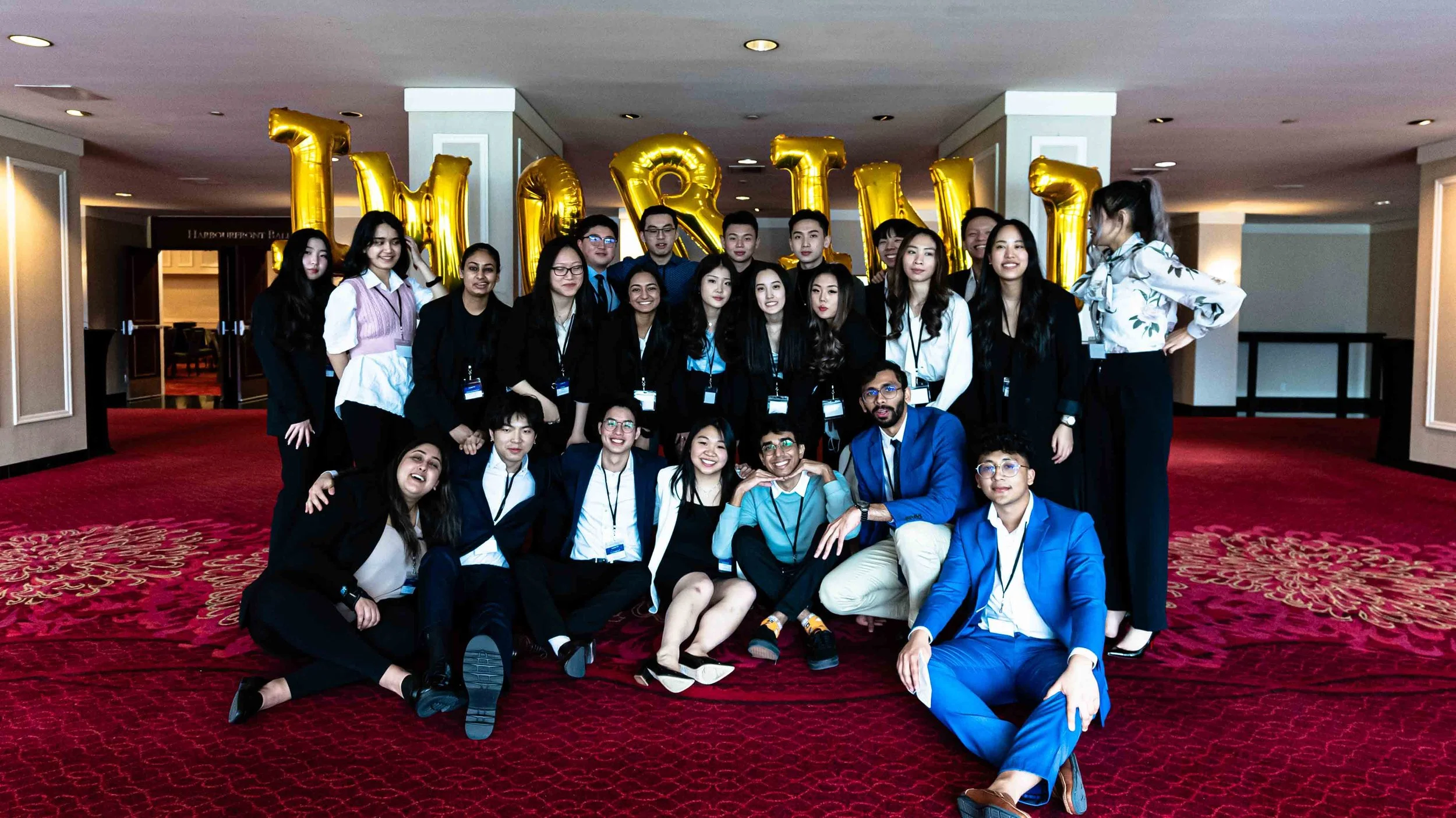 A group of young adults dressed in formal and semi-formal clothing posing for a photo in front of large gold balloons spelling out 'HIRE' at an indoor event.