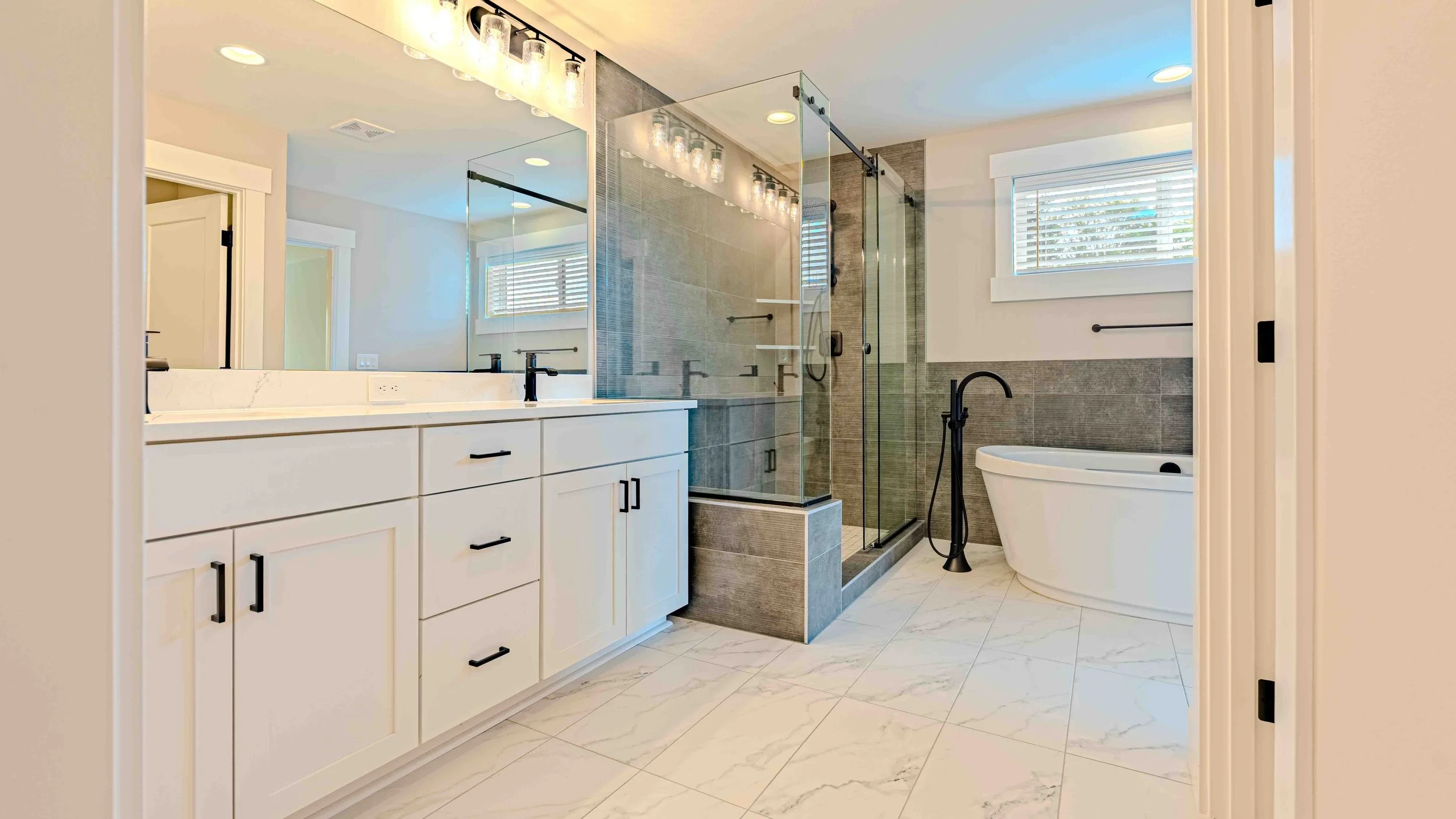 Modern bathroom with white vanity, black faucet, large mirror, glass enclosed shower with gray tiles, standalone bathtub, and windows with blinds.