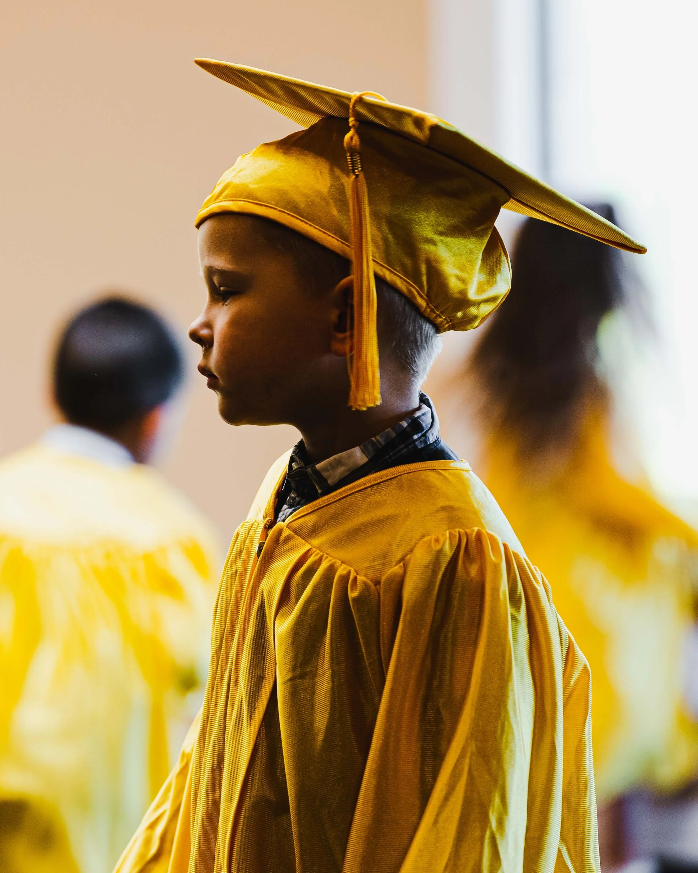 Young student wearing a yellow graduation cap and gown, standing in a classroom or ceremony setting.