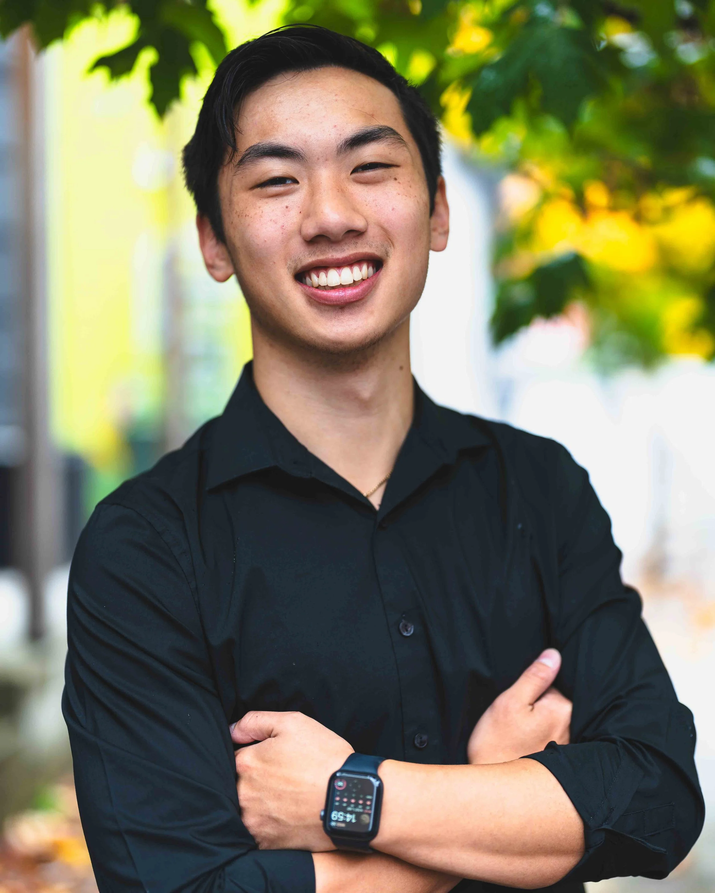 A young man with dark hair, wearing a black shirt and a smartwatch, smiling with arms crossed, standing outdoors with blurred green trees in the background.