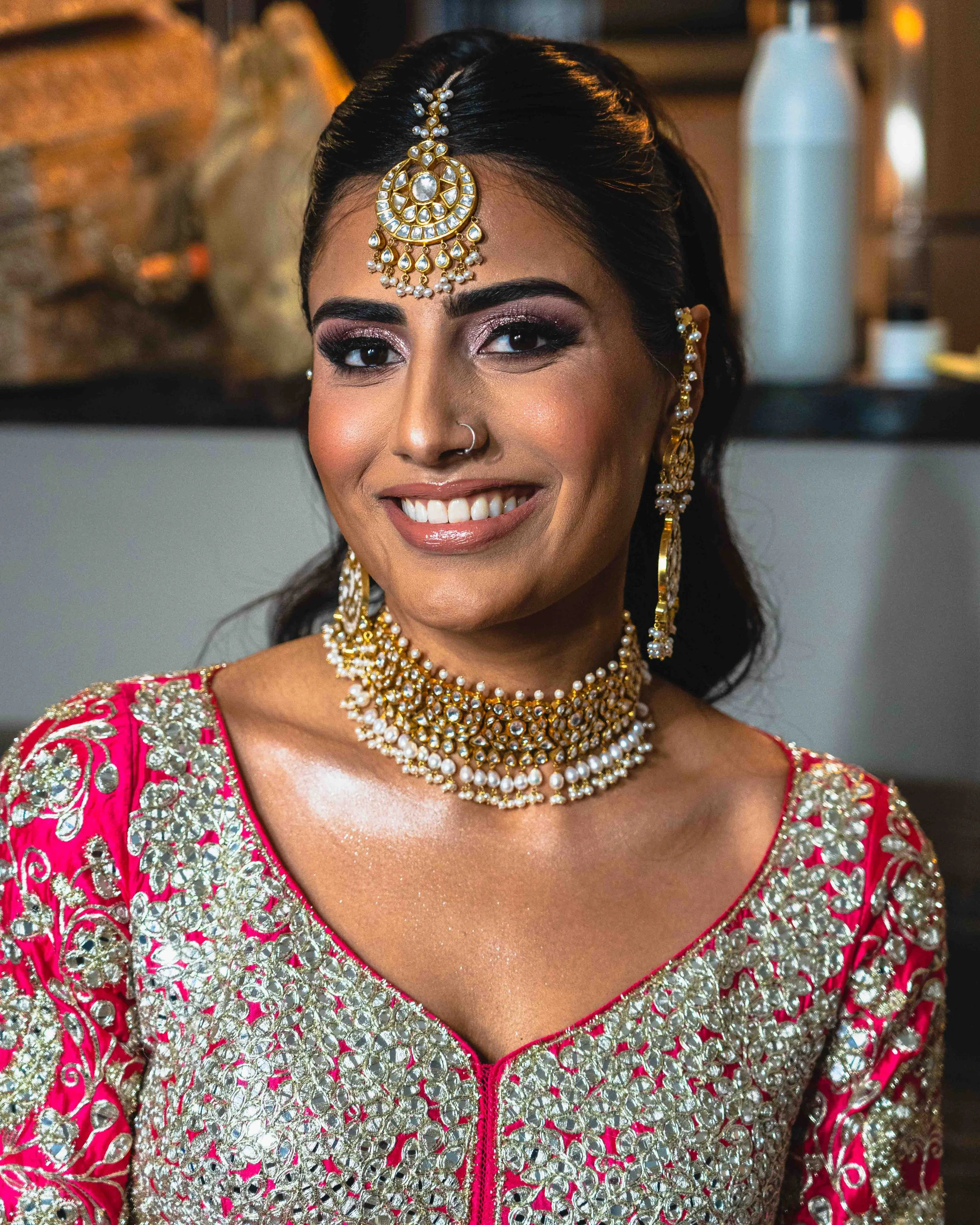 Close-up of a smiling woman dressed in traditional Indian attire, wearing elaborate gold and pearl jewelry, including a maang tikka, necklace, earrings, and a pink embroidered outfit.