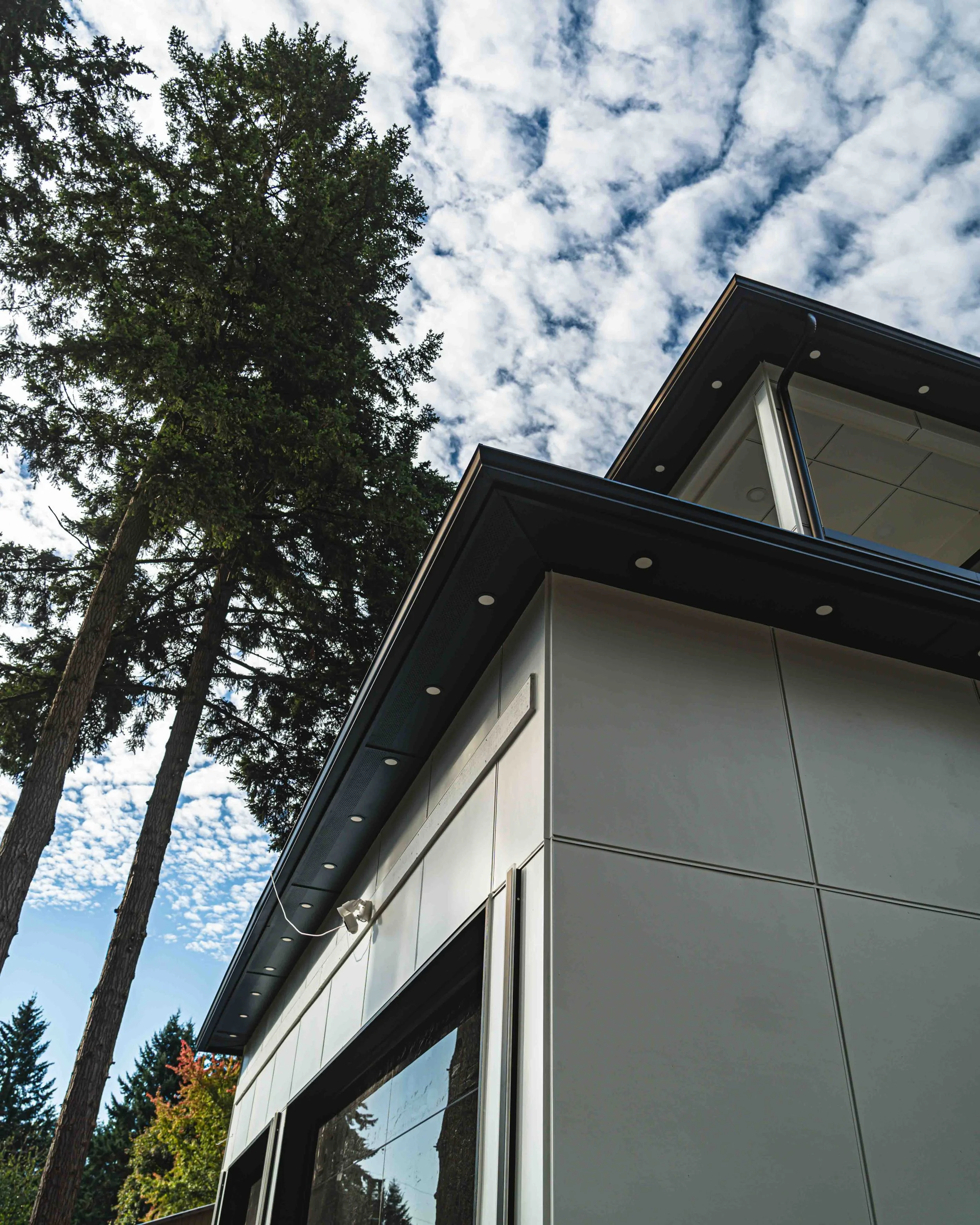 The image shows a close-up of the corner of a modern building with large glass windows and gray paneling. Tall pine trees are in the background with a partly cloudy sky overhead.