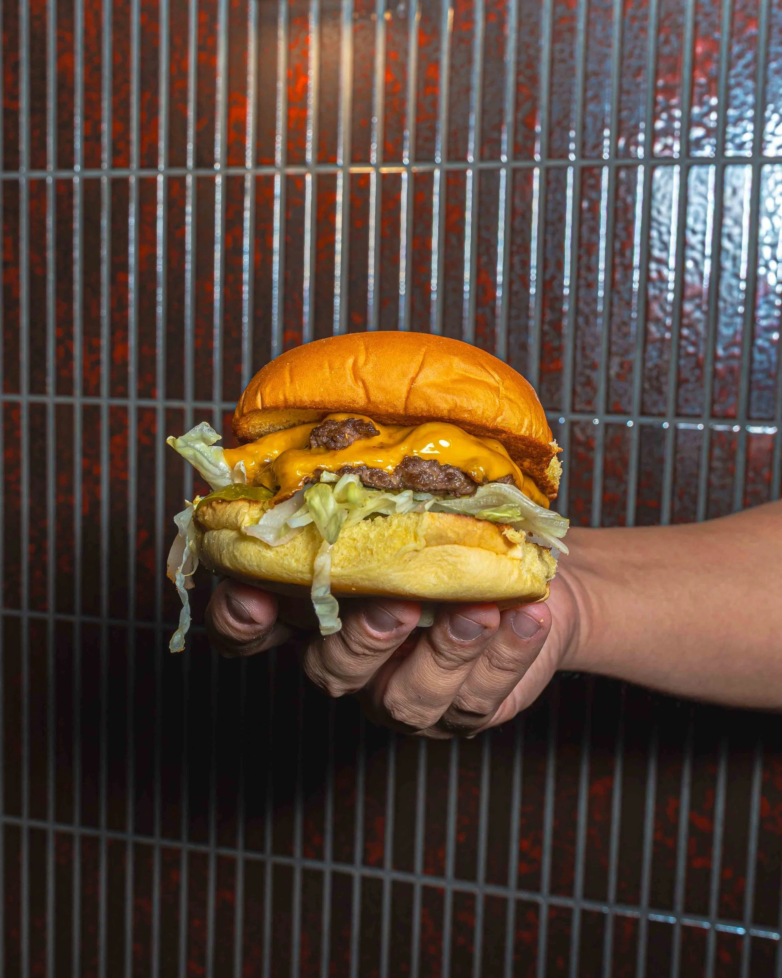 Hand holding a cheeseburger with lettuce, tomato, cheese, beef patty, and a bun against a metal grill background.