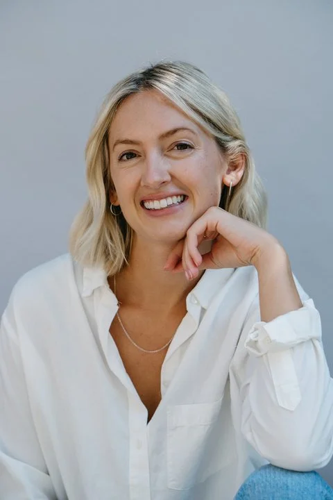 A woman with blonde hair smiling, wearing a white shirt and hoop earrings, resting her chin on her hand against a light background.