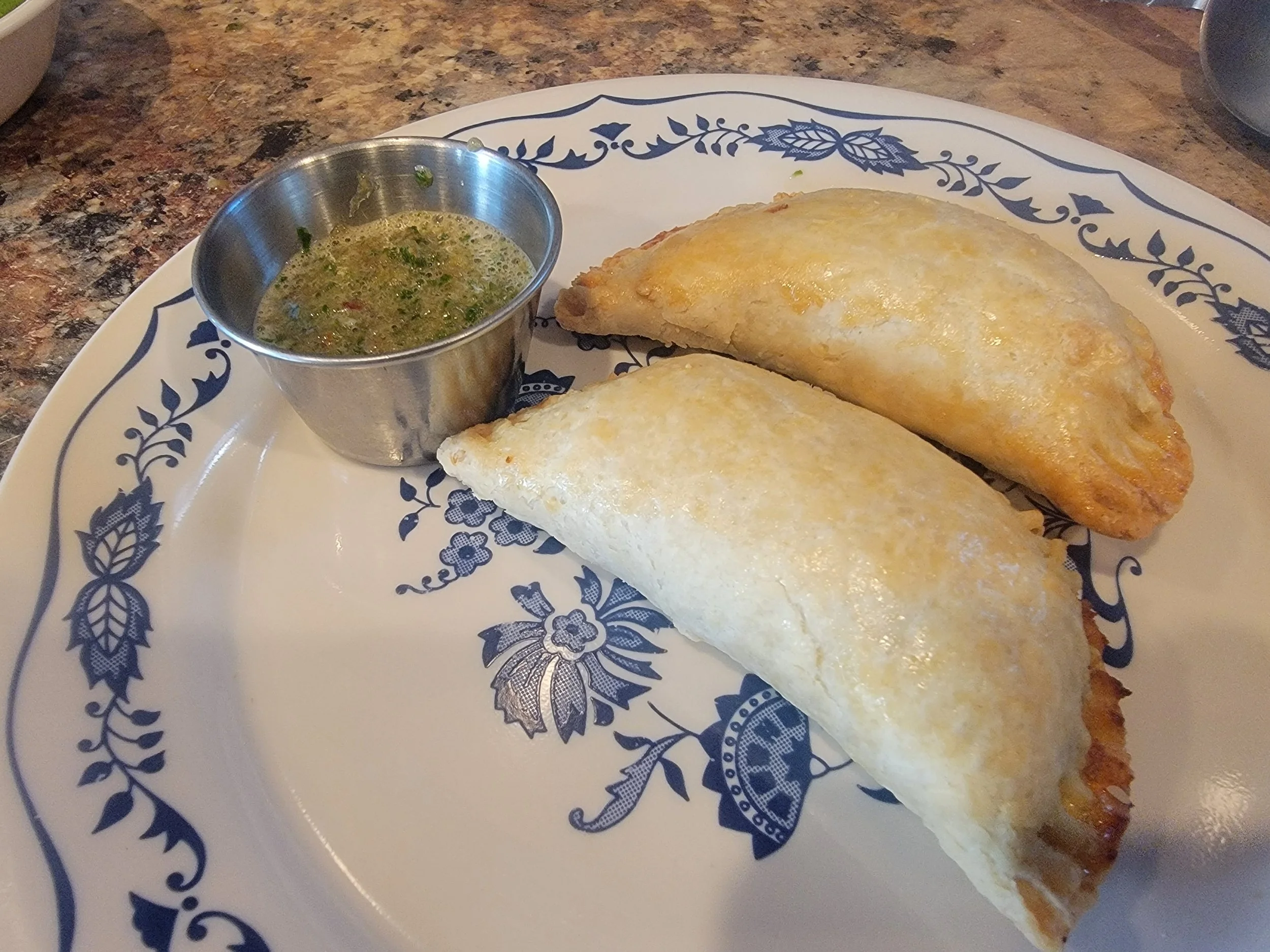 Two golden crescent-shaped pastries rest  atop a blue and white dish next to a small silver bowl of hot sauce.