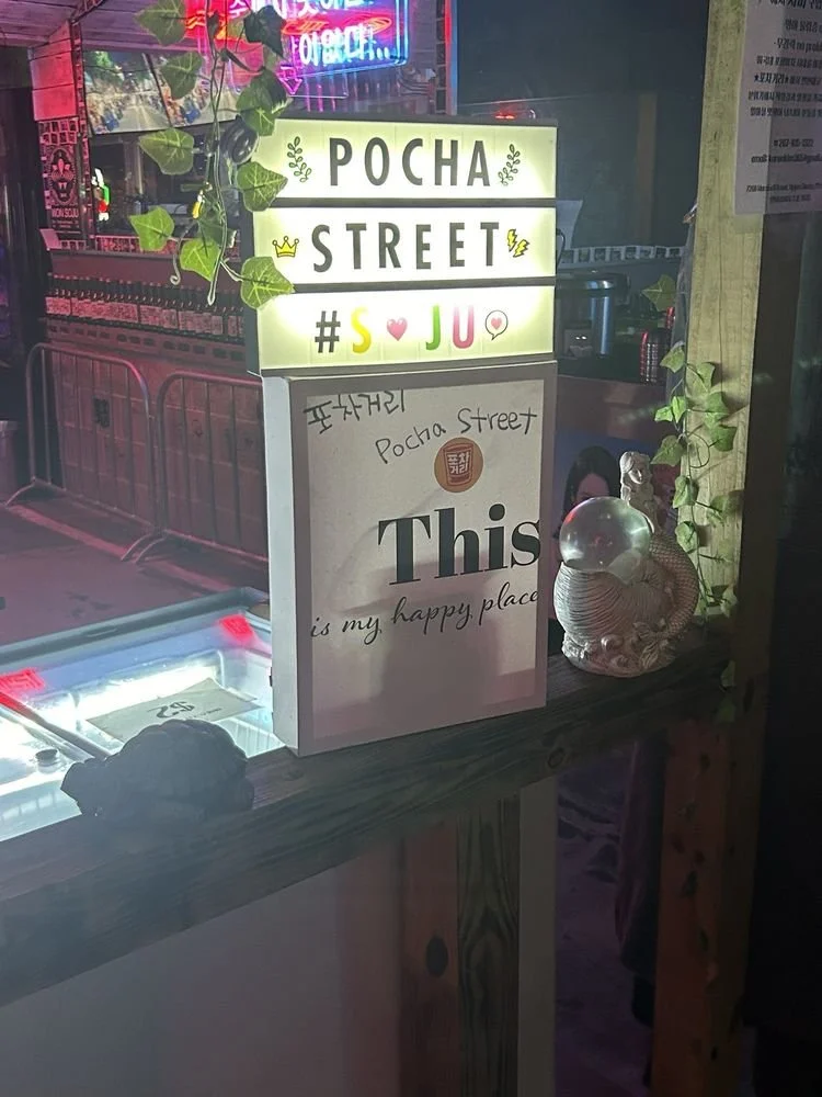 Illuminated signboard reading 'Pocha Street' with colorful emojis and text, next to a decorative snail sculpture and a small glass orb on a wooden surface at night, with city lights in the background.