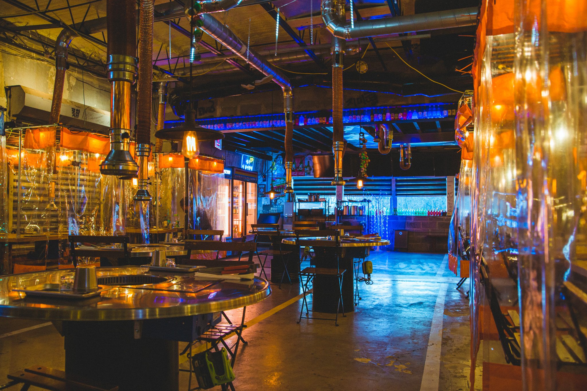 Inside a vibrant restaurant or bar with hanging industrial-style lamps, colorful lighting, and metallic decor. Tables and chairs are set up, and the space features visible pipes and ductwork on the ceiling, creating a modern urban ambiance.