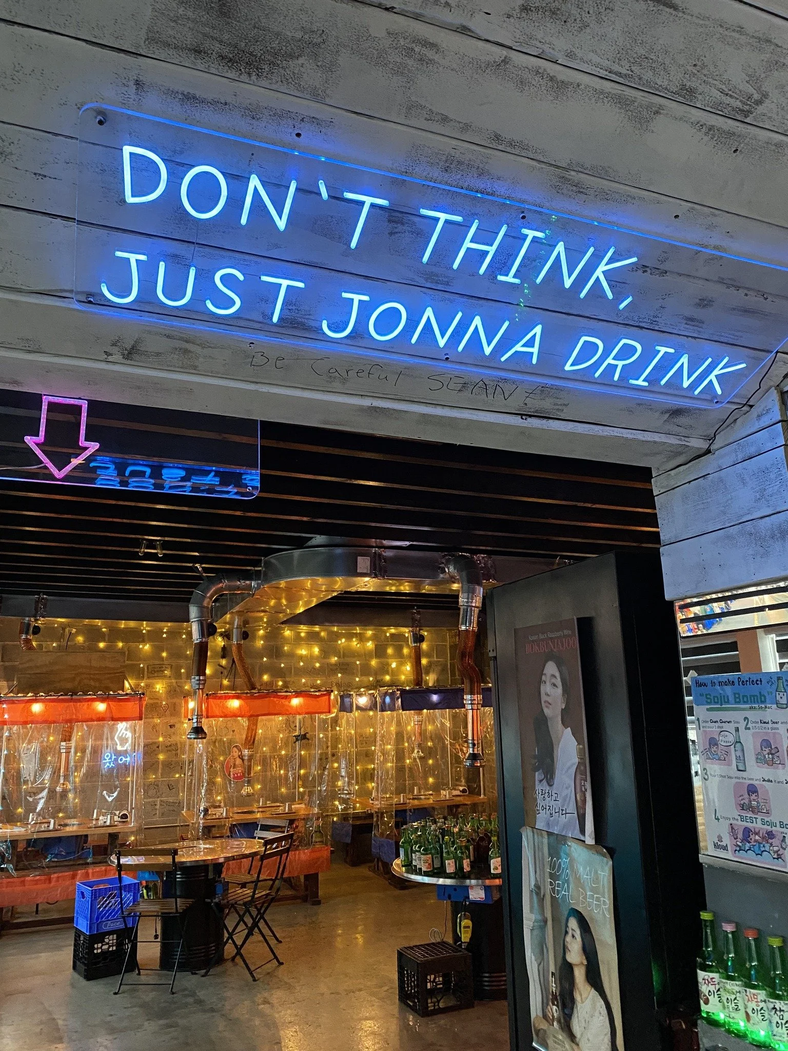 Neon sign with blue text reading "DON'T THINK, JUST JONNA DRINK" above an interior of a bar or restaurant, illuminated with warm string lights, glasses, bottles, and posters.