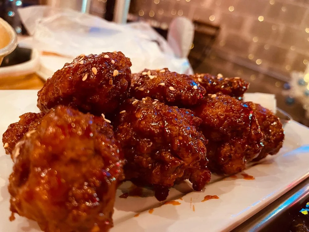 Close-up of a plate of crispy glazed chicken wings sprinkled with sesame seeds, placed on a white parchment paper on a serving tray.