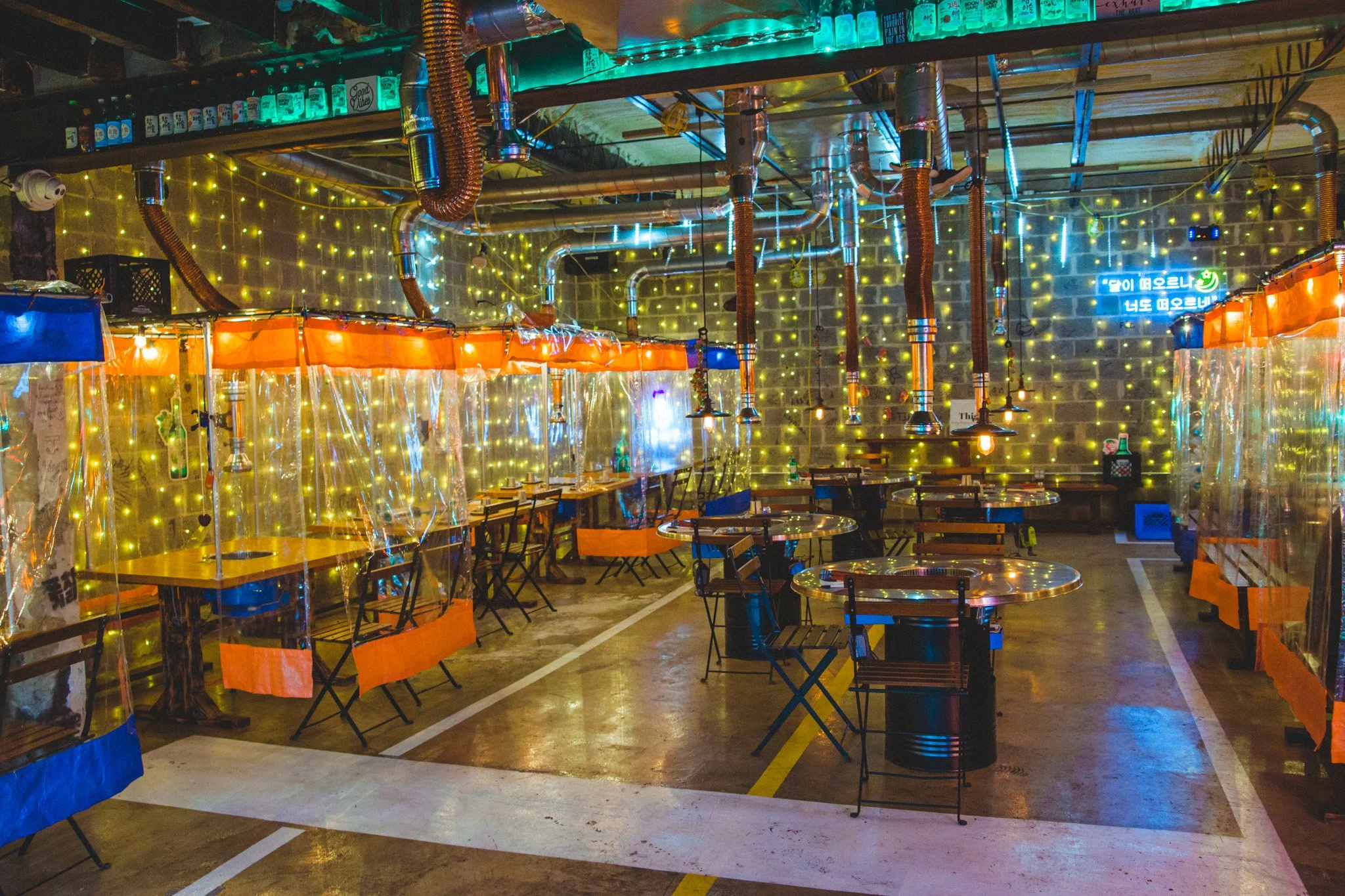 An empty indoor restaurant or bar decorated with warm string lights, neon signs, and hanging pipes for a cozy atmosphere. There are several tables with chairs, some with red and blue curtains or barriers for partition. The space has industrial elements like exposed ducts and pipes, and the overall lighting is colorful and dim.