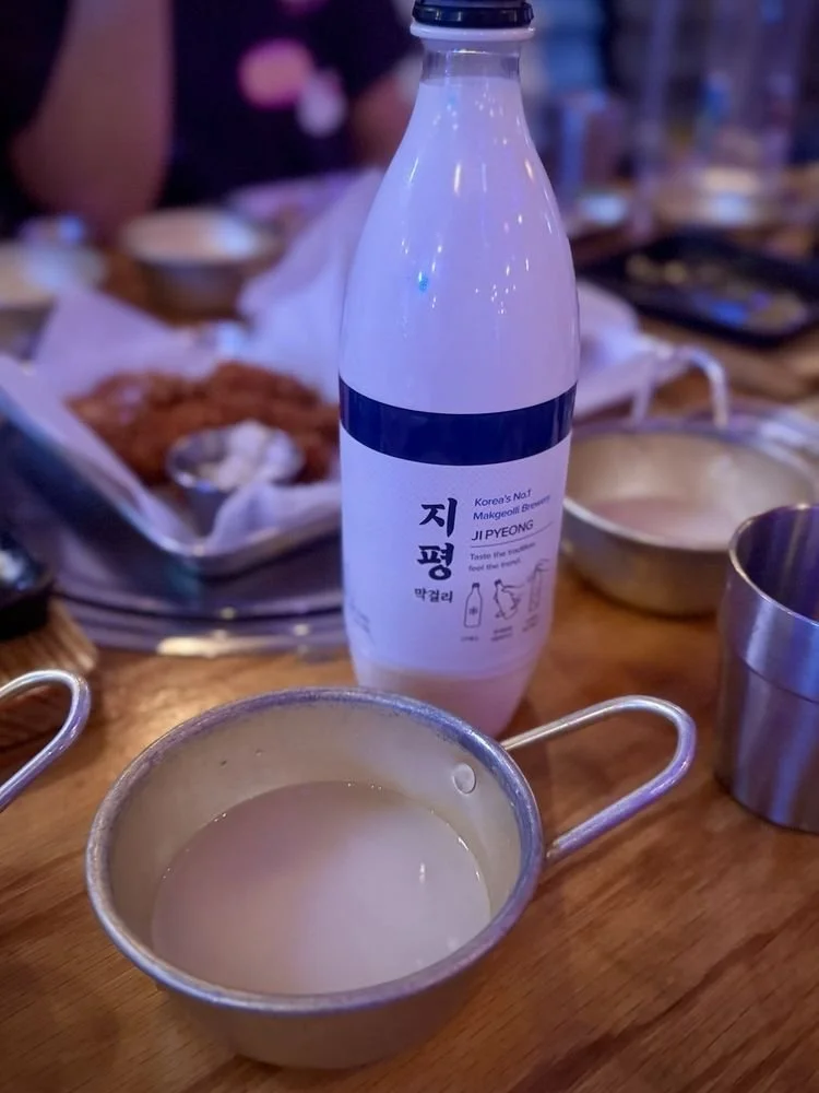 A bottle of Korean rice wine (makgeolli) on a wooden table with a metal cup filled with the drink in front of it, alongside dishes of Korean food in the background.