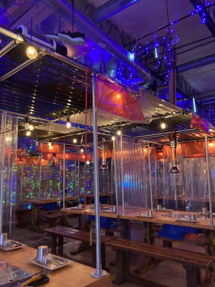 Decorated indoor restaurant with string lights, colorful string lights on walls, wooden tables with metal plates and cups, clear plastic dividers, and exposed ceiling pipes with string lights and decorations.