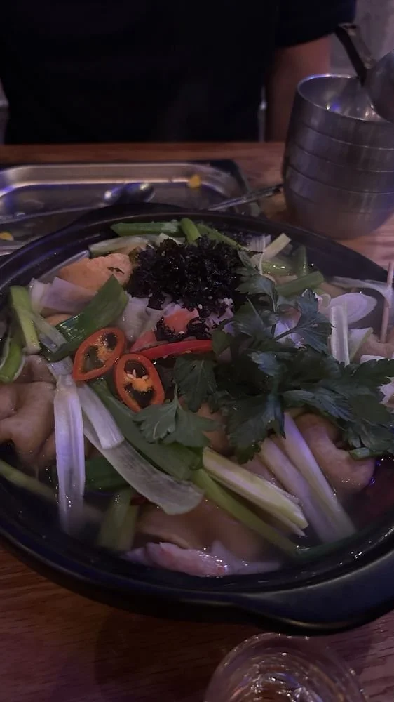 A bowl of Asian hot pot with vegetables, sliced chili peppers, and herbs on a wooden table. There are metal bowls and utensils nearby.