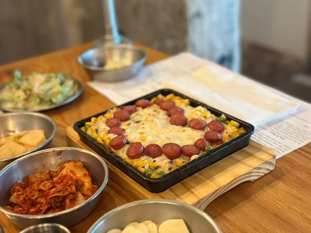 A cast iron skillet of baked casserole topped with slices of sausage and melted cheese, surrounded by small metal bowls of side dishes on a wooden table.