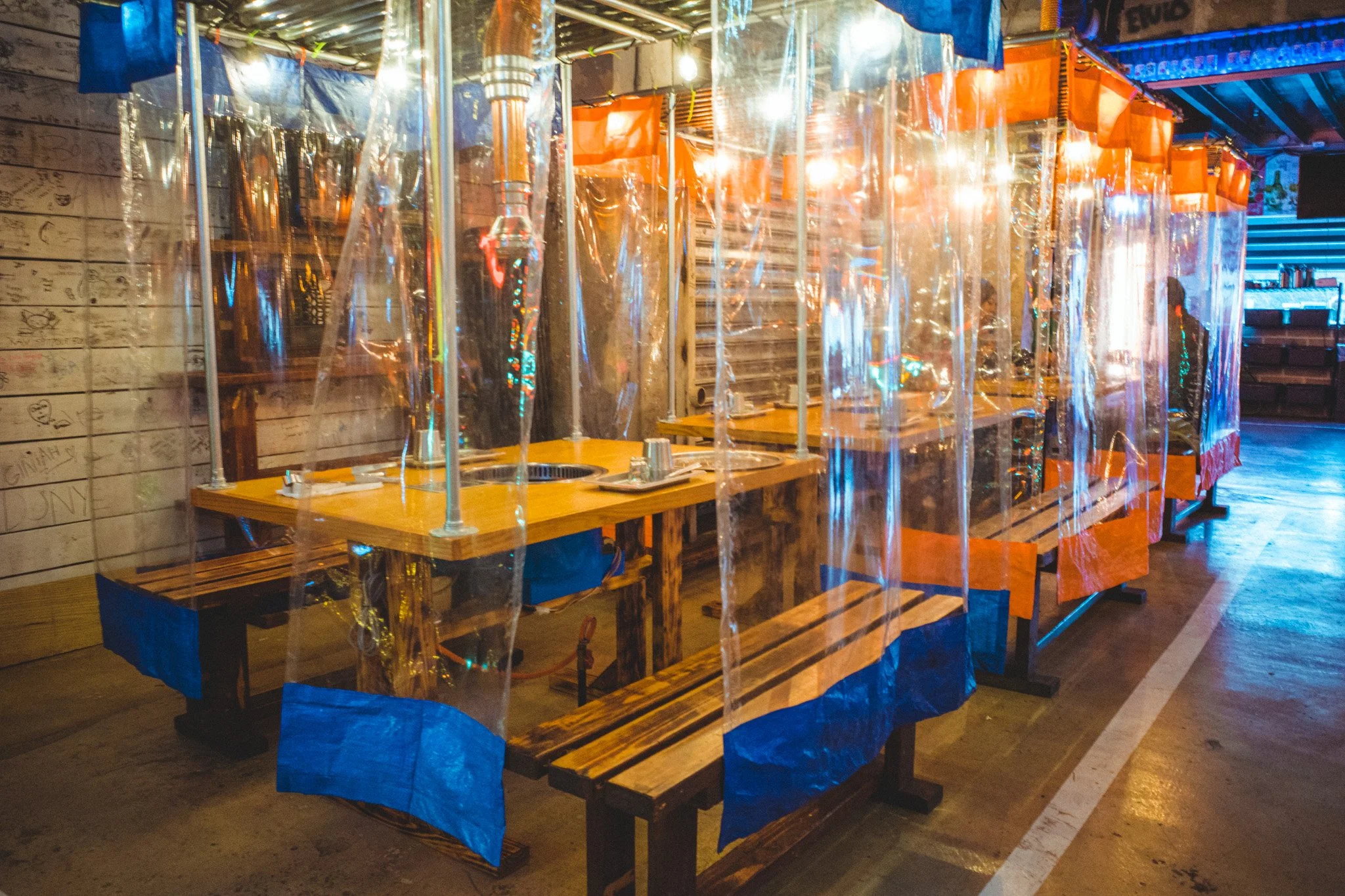 Outdoor seating area with wooden tables and benches separated by clear plastic dividers, possibly for social distancing, illuminated by warm lighting, with a decorative wall on the left and a bar or counter area in the back right.