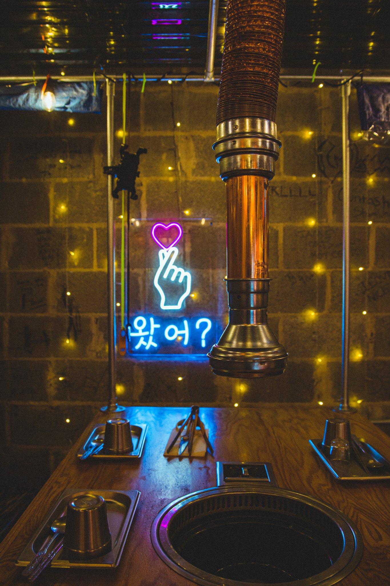 A restaurant table with built-in grill and metal utensils, set against a wall decorated with yellow fairy lights and a neon sign depicting a hand with a finger pointing to a pink heart, with Korean characters below.