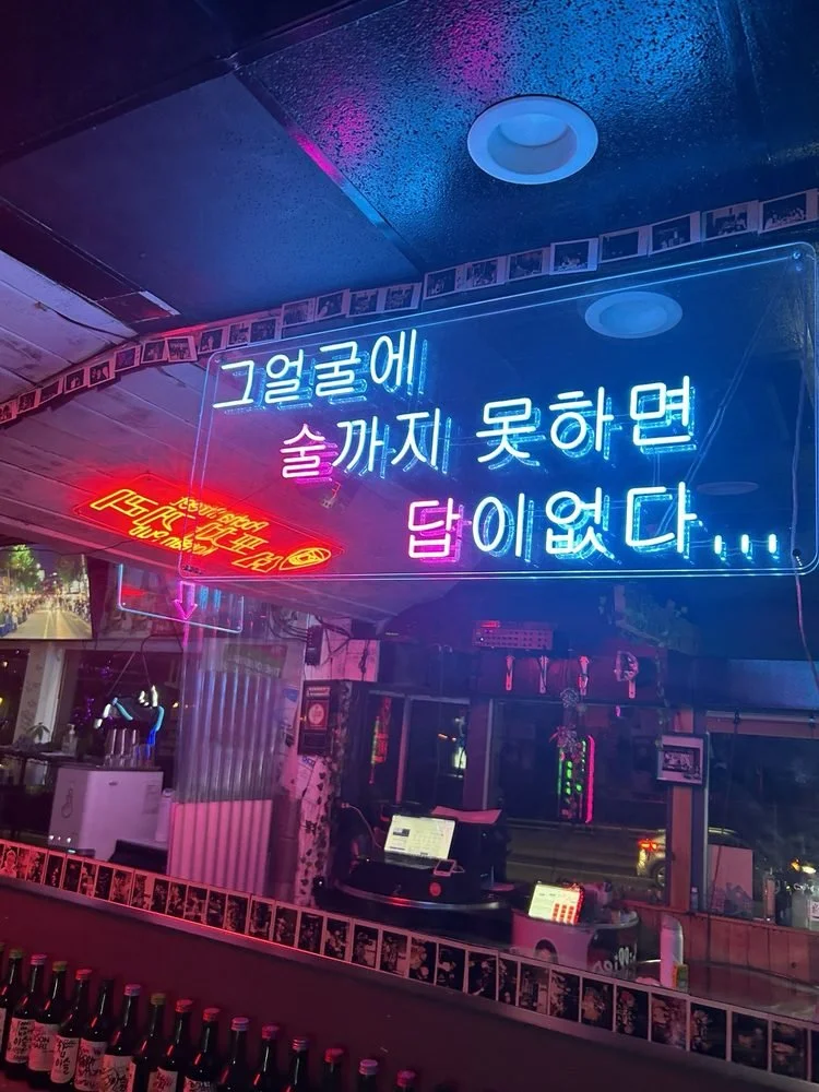 Neon sign in Korean with blue, pink, and red lights inside a bar or restaurant, reflecting on the glass window.
