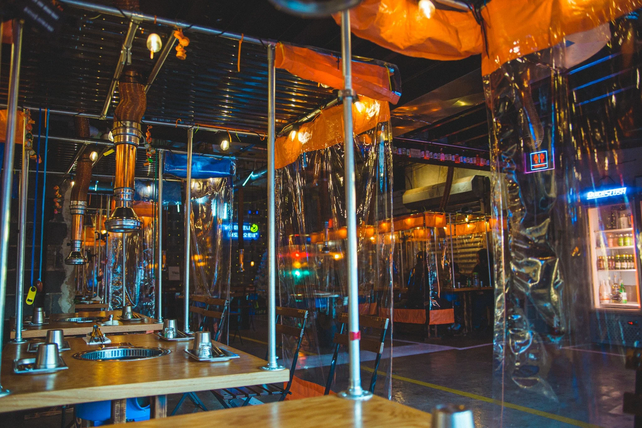Indoor restaurant with wooden tables, metal poles, and plastic dividers, illuminated by colorful neon lights