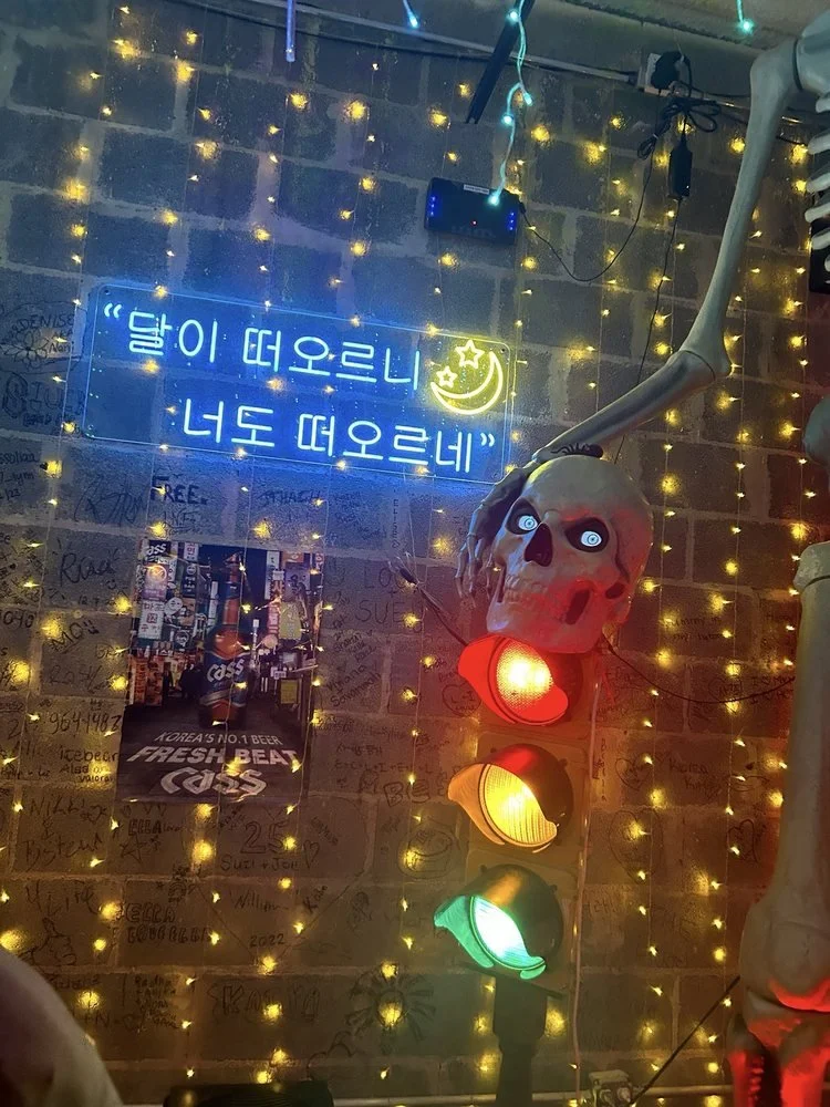 Decorative scene with a skull mask, string lights, and a neon sign with Korean text and a crescent moon with a star, along with a traffic light and posters on a brick wall.