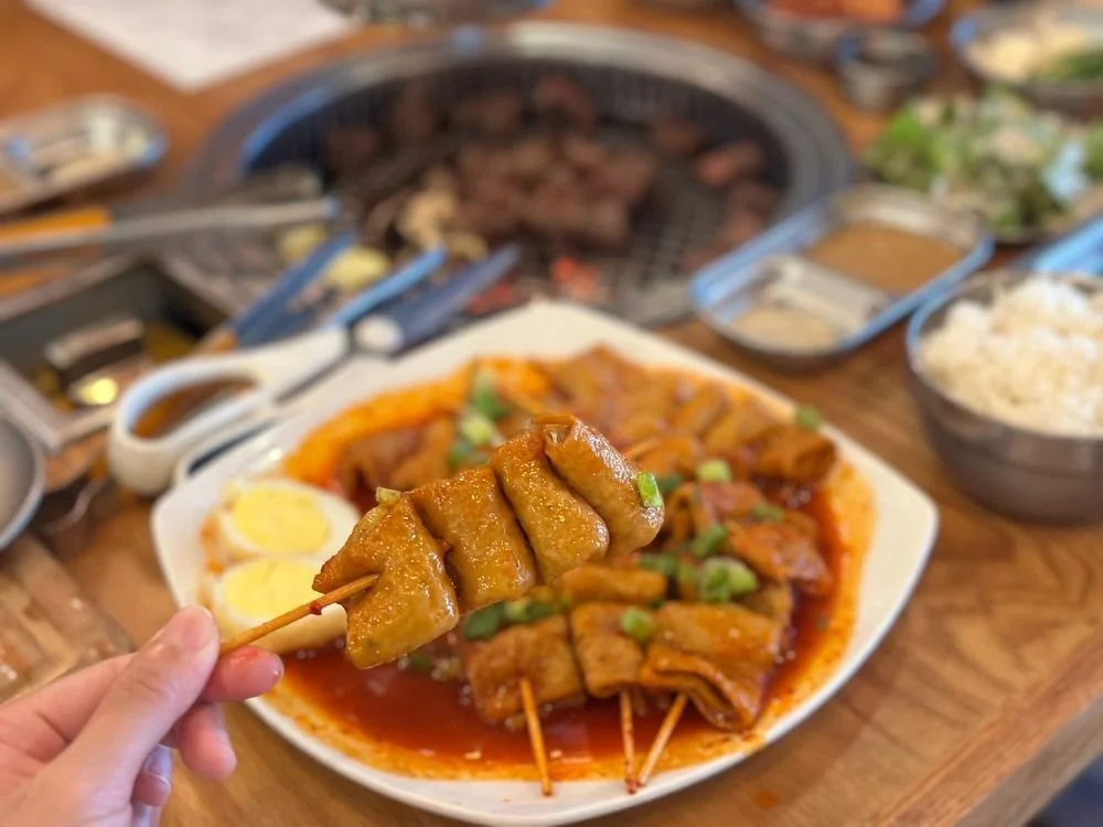 Close-up of skewered grilled meat and vegetables on a plate with rice and various side dishes in the background at a Korean barbecue restaurant.