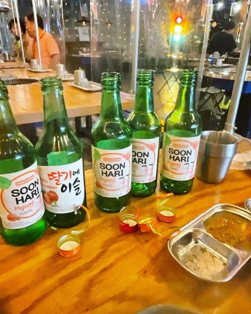 Four green glass bottles of Soon Hari yogurt drink on a wooden table, with several bottle caps and a stainless steel tray with food. Background shows a restaurant setting with people sitting at tables and a traffic light visible outside.