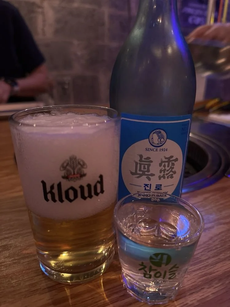 A glass of beer with foam, a bottle of Korean soju, and a small glass of clear liquid on a wooden table in a dimly lit setting.