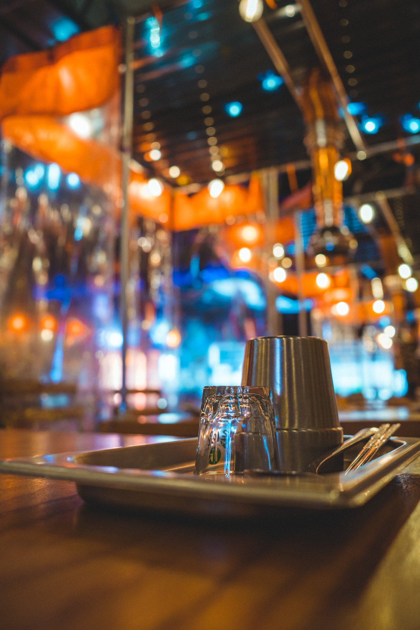 A restaurant table setting with upside-down metal cups, a glass, and cutlery on a tray, with blurred colorful lights and interior in the background.