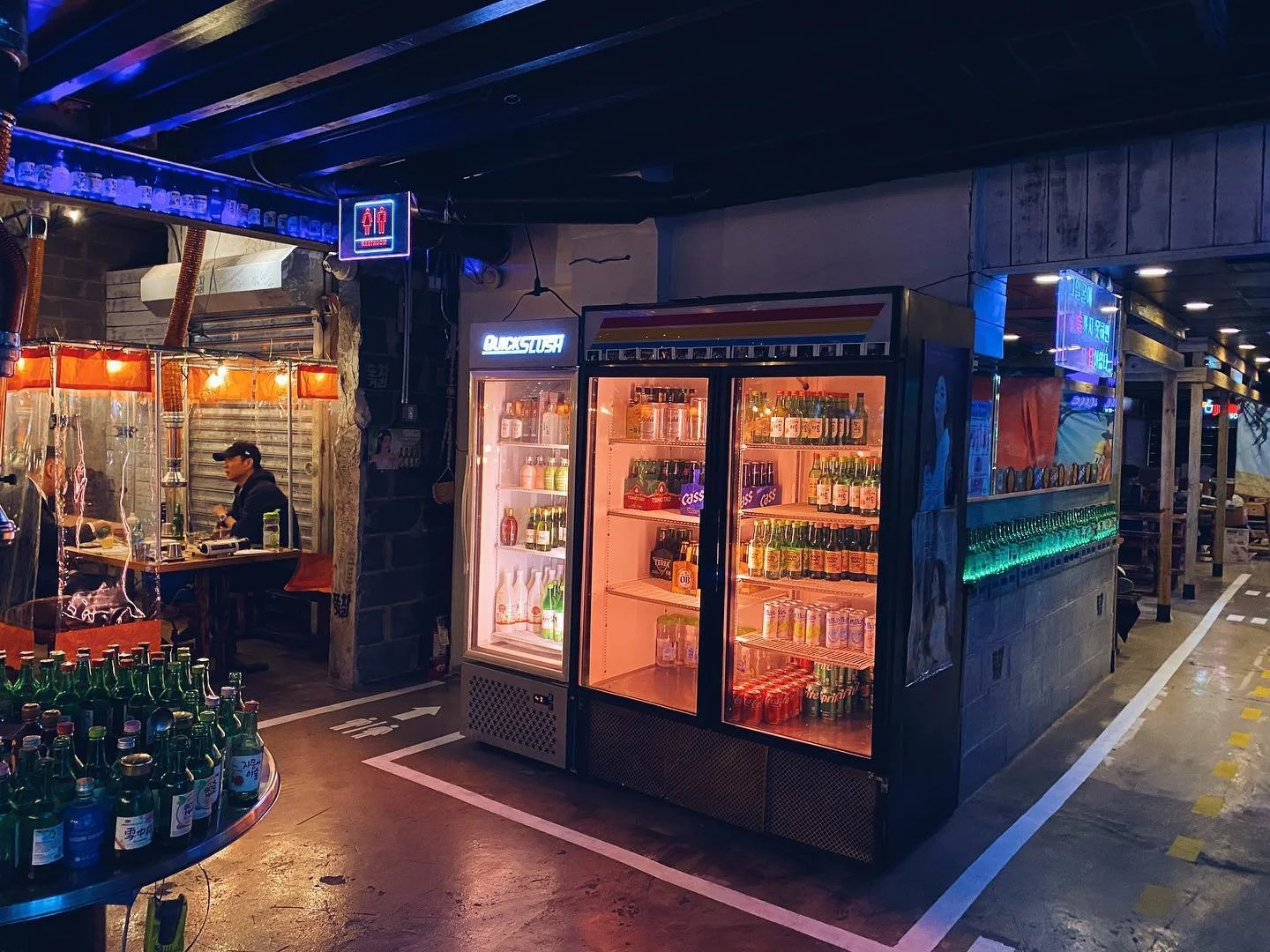 A nighttime scene in a bar or restaurant with a drink refrigerator filled with various bottles of alcoholic beverages, a neon sign, and a few customers sitting at a table to the left. The setting has a dimly lit, cozy atmosphere with colorful neon li
