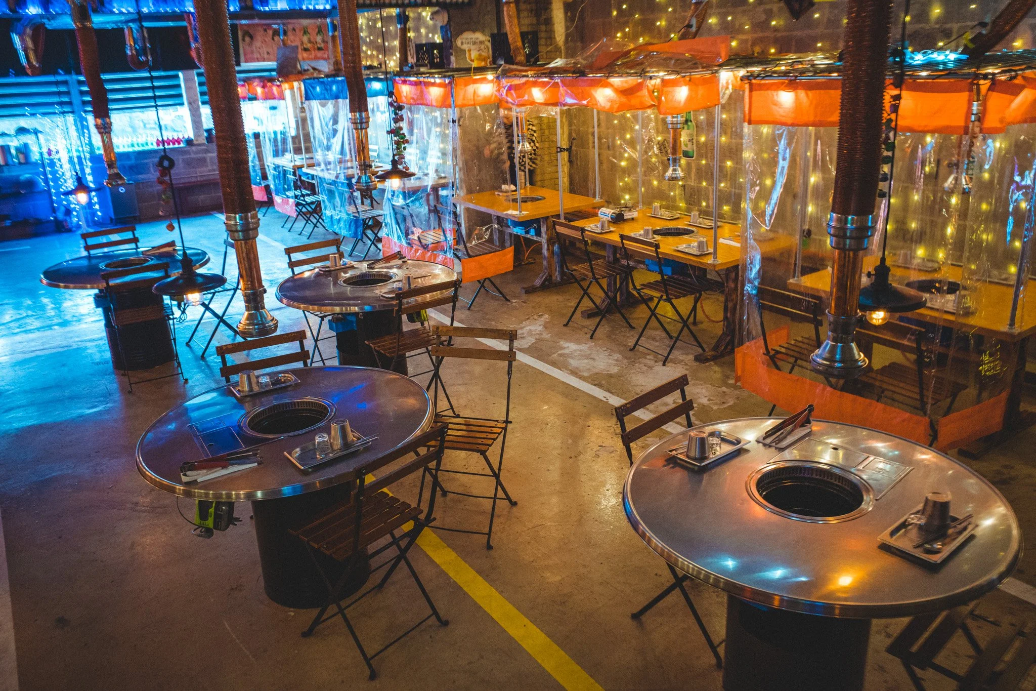 Empty restaurant with round and rectangular tables featuring built-in grills, arranged in a large open space with colorful string lights, plastic dividers, and hanging exhaust vents.