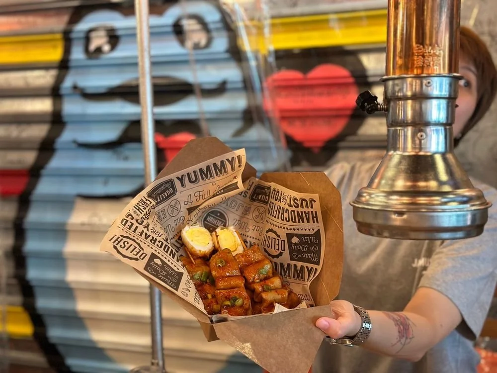 Person holding a paper boat with food, including sausages and boiled eggs, in front of a graffiti painted shutter with a face and heart.