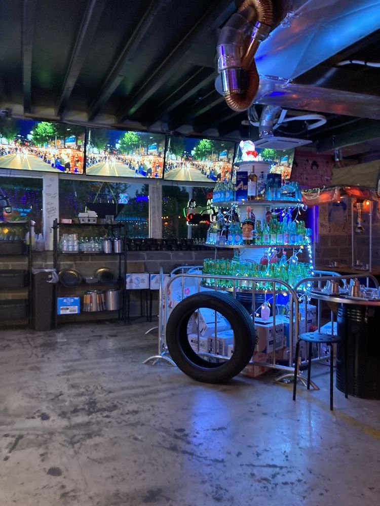 Indoor bar area with shelves stocked with bottles and glasses illuminated by blue lights, a TV displaying a city scene, a tire hanging on a cart, and a background of a dimly lit room with tables and chairs.