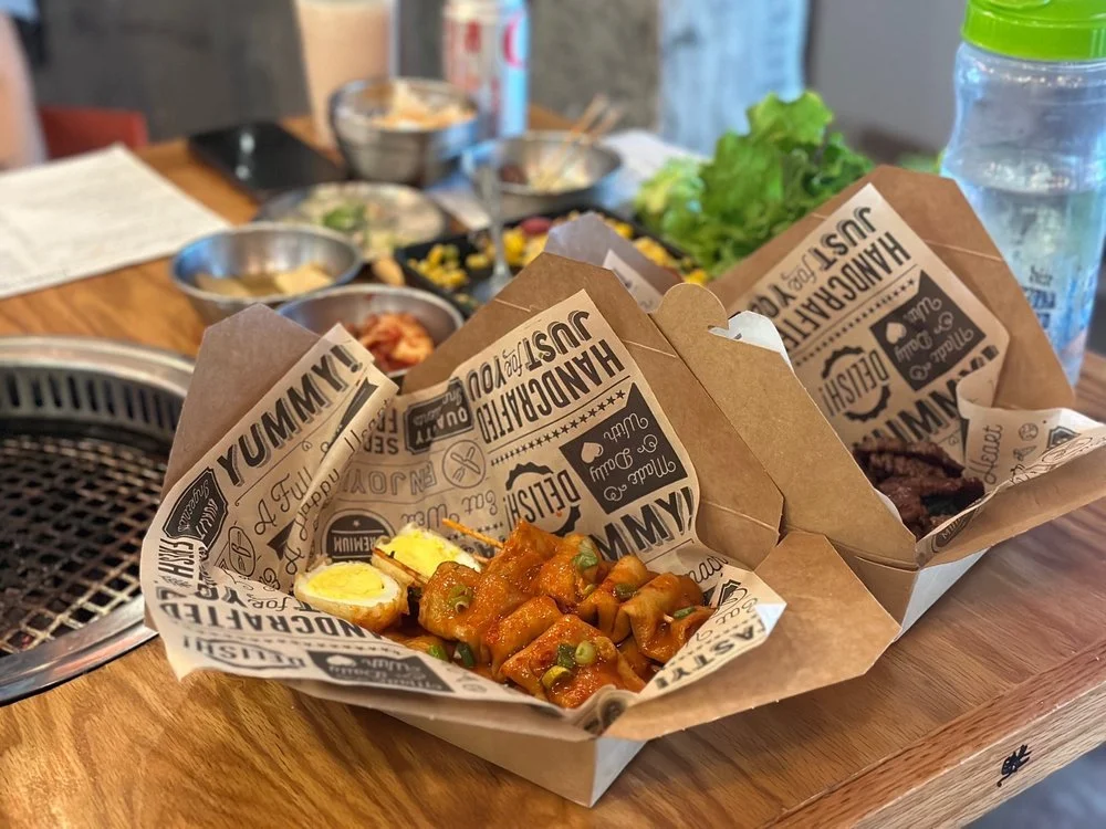 Three takeout boxes with food on a wooden table, containing drinks and various side dishes.