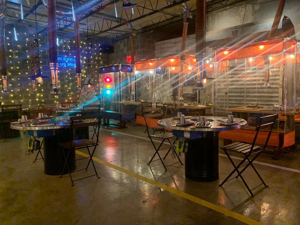 Restaurant with round tables set with utensils, chairs, and plastic barriers, decorated with string lights and illuminated signs, suggesting a modern dining space with safety protocols.
