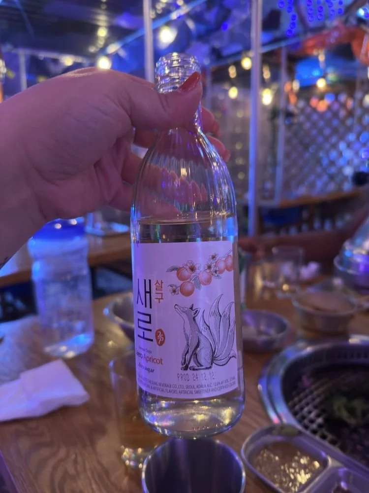 A hand holding a clear plastic bottle of water with a label featuring a fox and apricots, set against a background of a restaurant or bar with blue and purple lights and a grill.