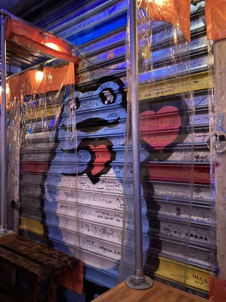 Wall with graffiti of two profiles and a heart between them, covered with handwritten messages and signatures, with metal poles and orange plastic sheeting.
