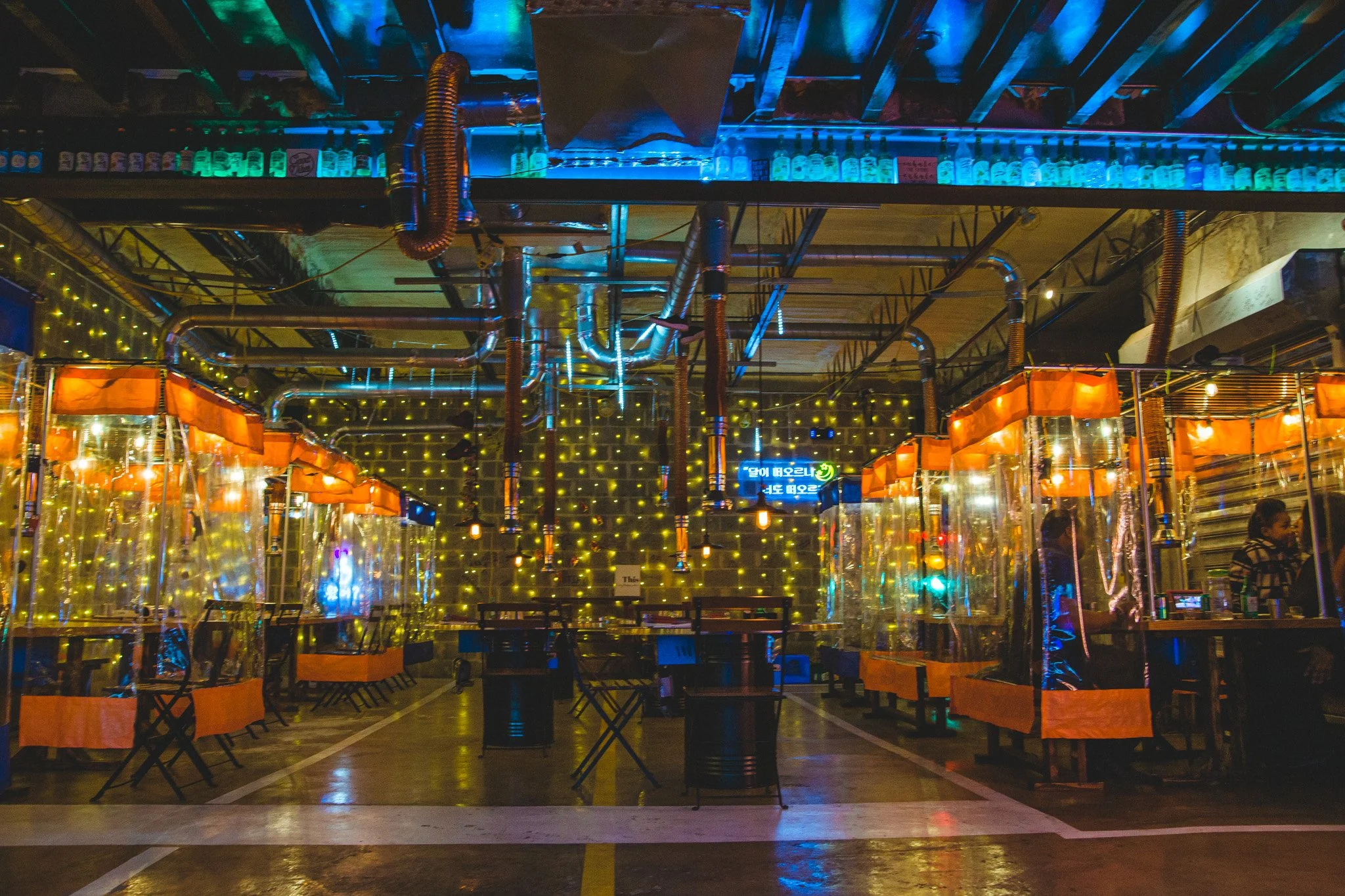 Indoor party venue with transparent plastic booths, string lights on the wall, and metal tables and chairs. Colorful lighting and exposed ceiling pipes are visible.