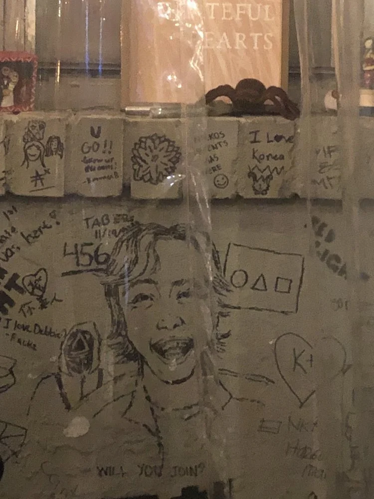 A wall covered with various drawings, doodles, and writings, including a large portrait of a smiling woman, geometric shapes, hearts, and text such as "WILL YOU JOIN?" and "I love Korea."